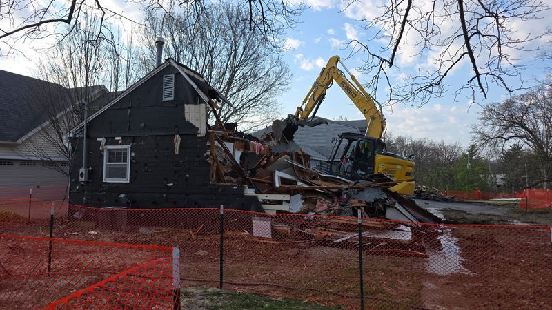 Yellow excavator demolishing a dark house behind orange safety fencing on a muddy lot.