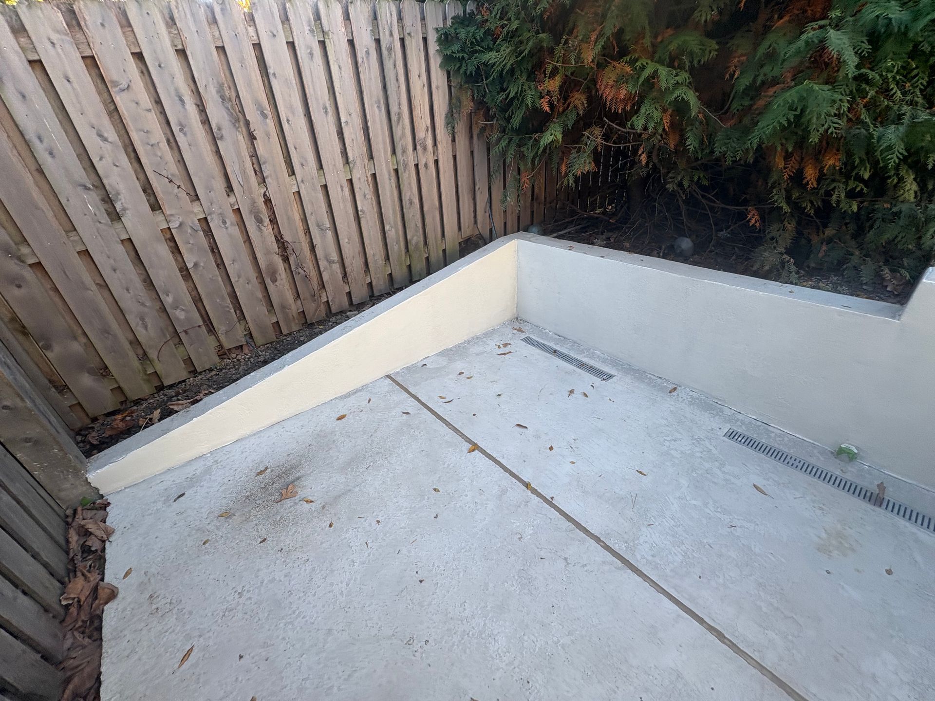 Empty concrete patio corner with wooden fence and shrubs nearby.