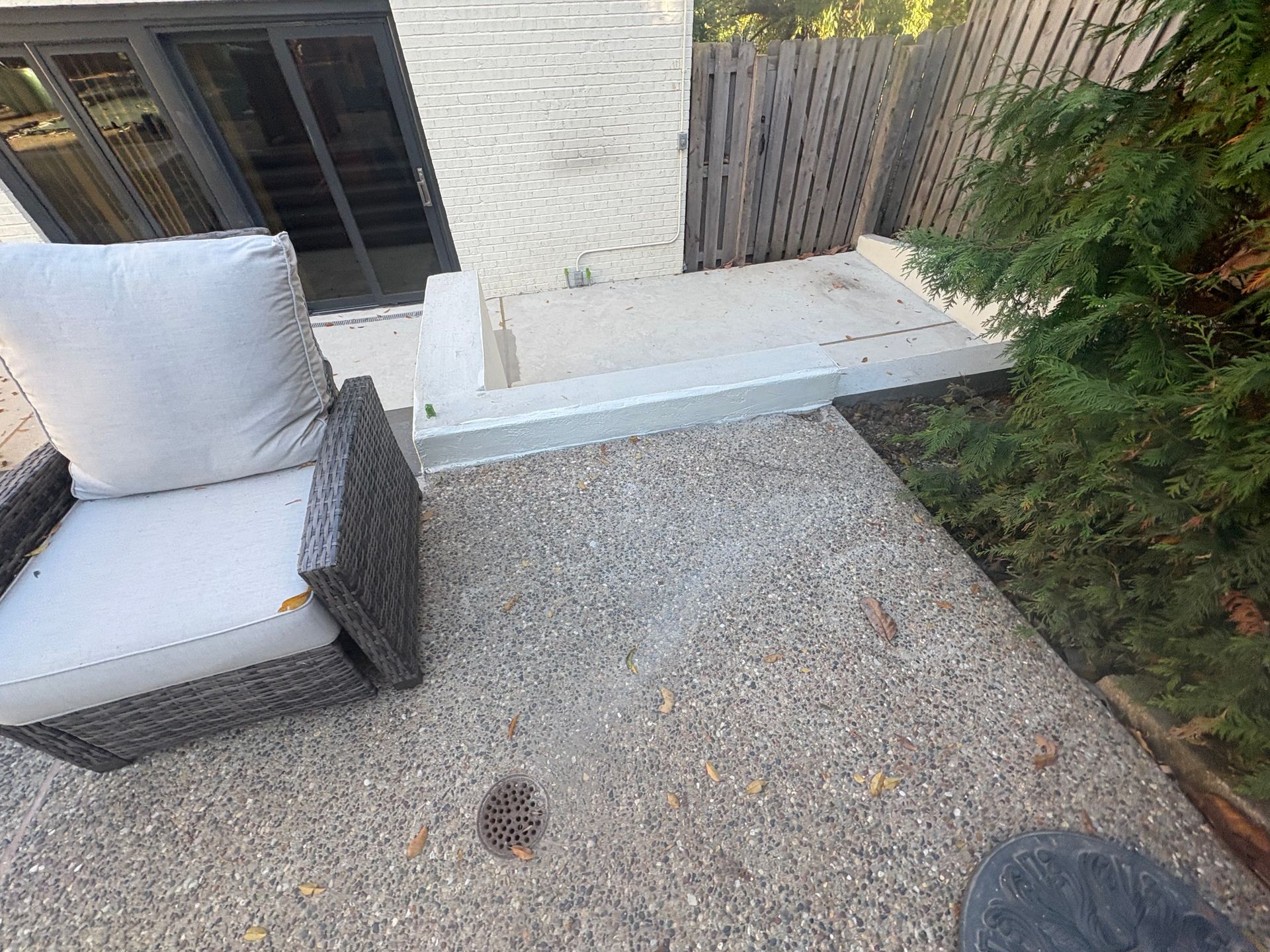 Small gravel patio with wicker chair, white cushions, concrete bench, and surrounding greenery.