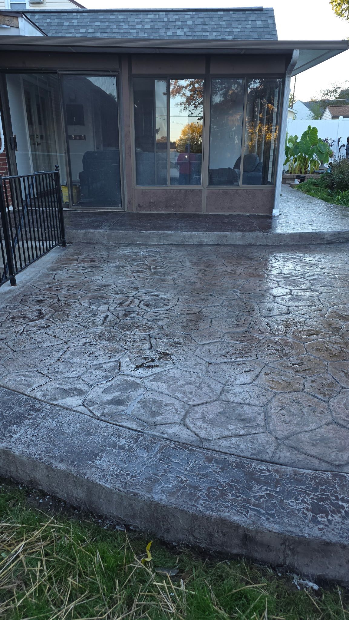 Stone patio leading to a screened front porch on a house, with grass at the edge and a black railing on the left.