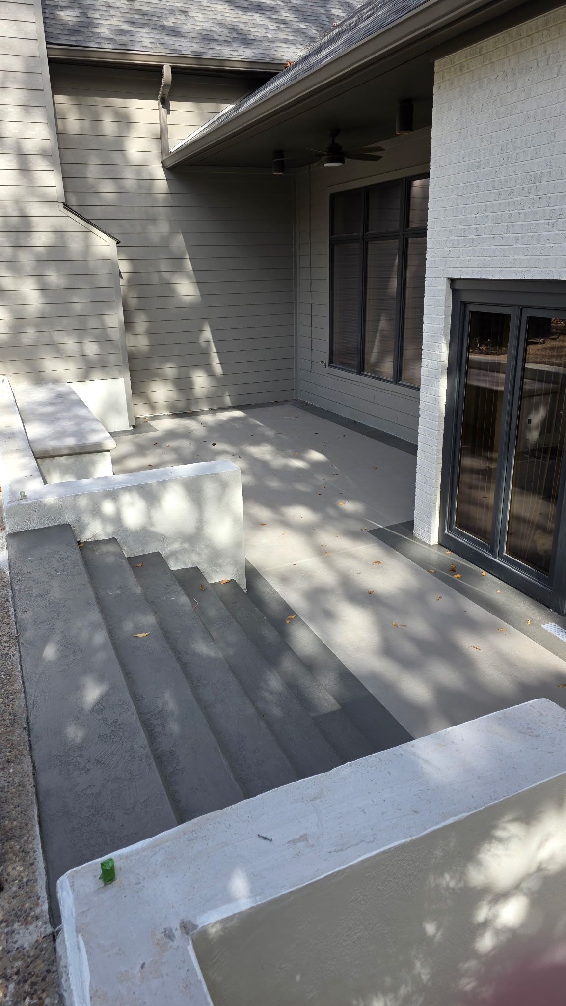 Covered front patio with gray pavers, white siding, and a glass door in sunlight and shadow.