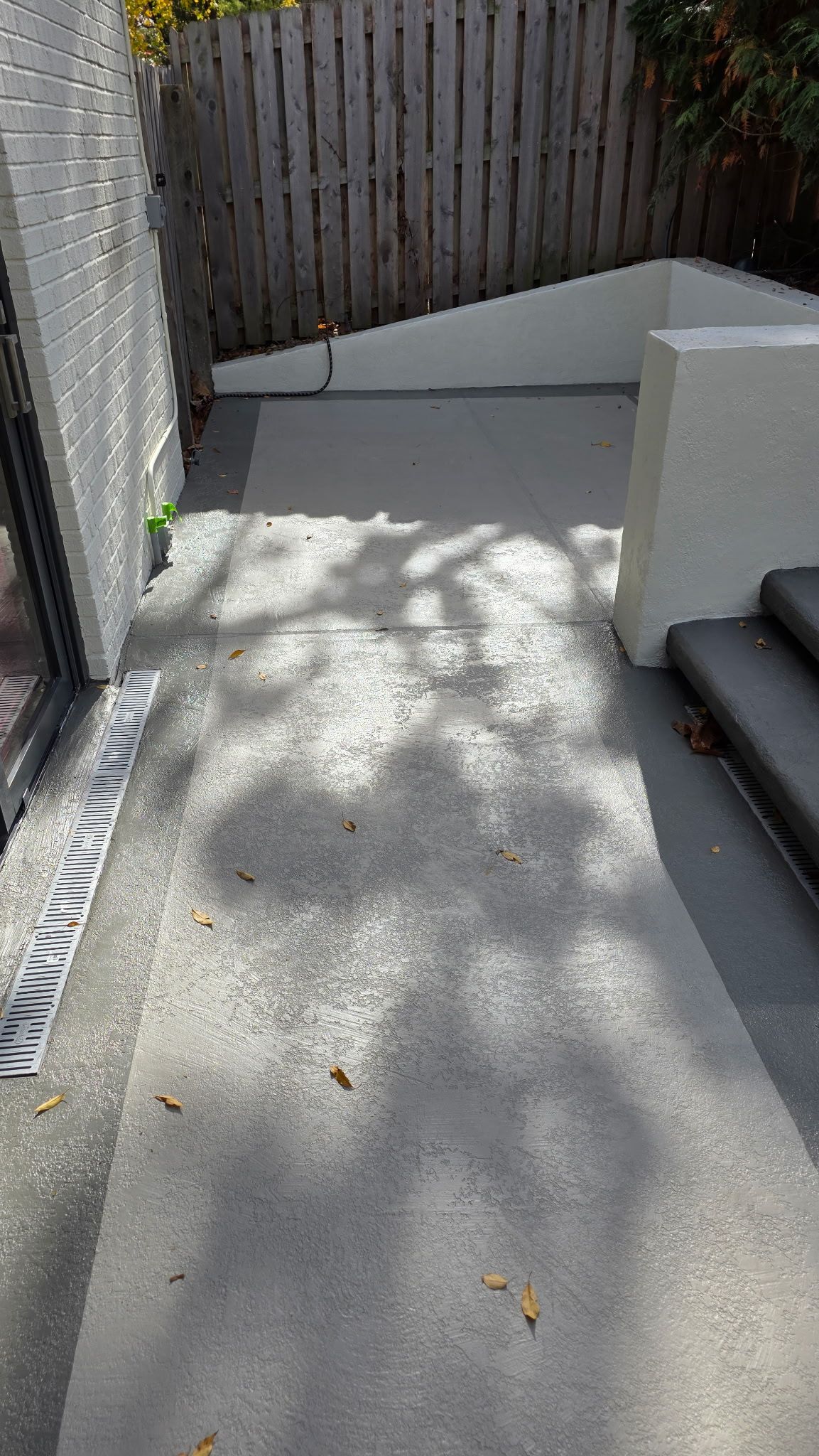 Concrete patio path beside stairs and a fence, with dappled sunlight and a small planter wall.