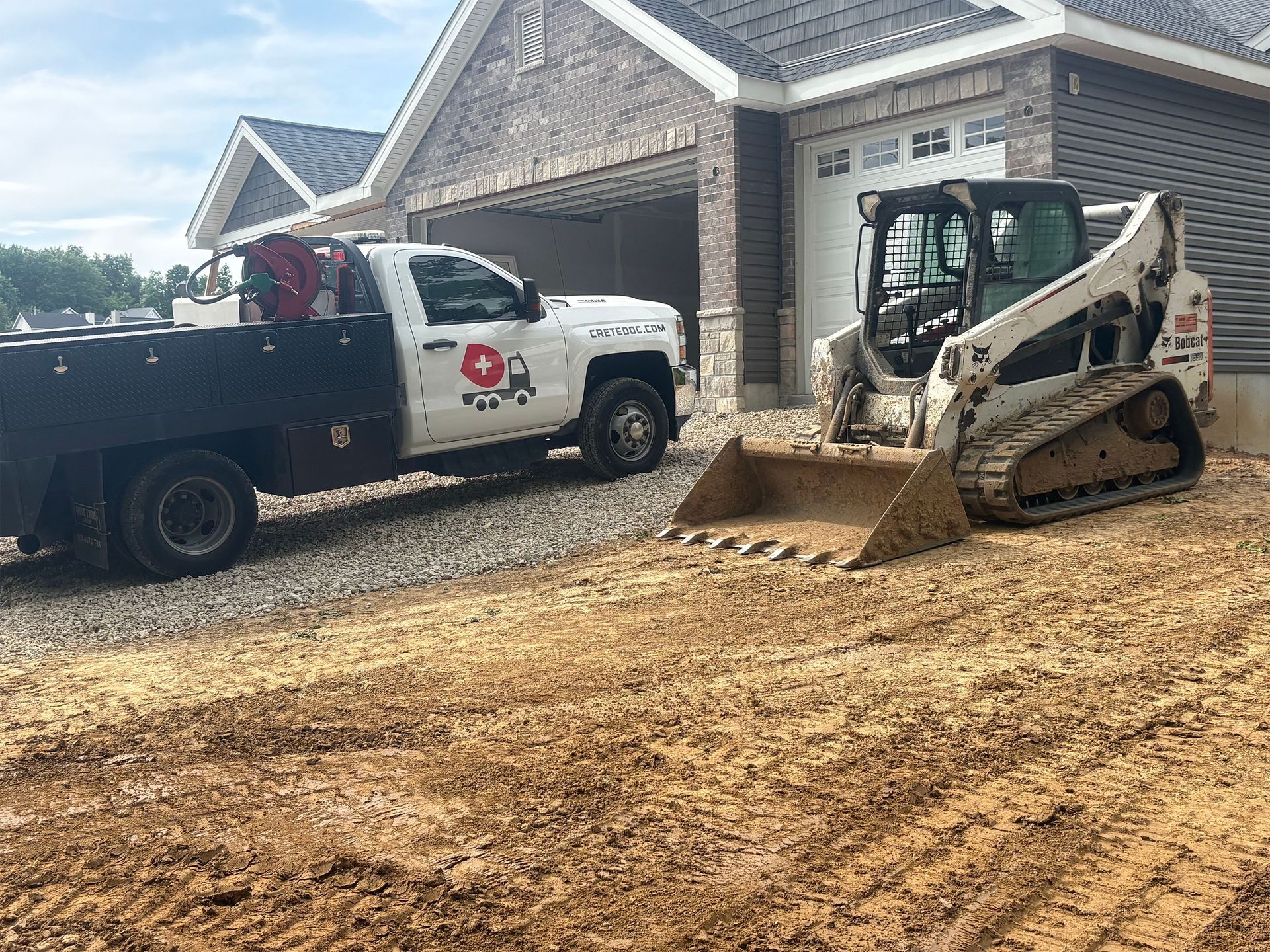Construction vehicles outside a house, with a red cross truck and a skid steer unloading dirt in front of the garage.