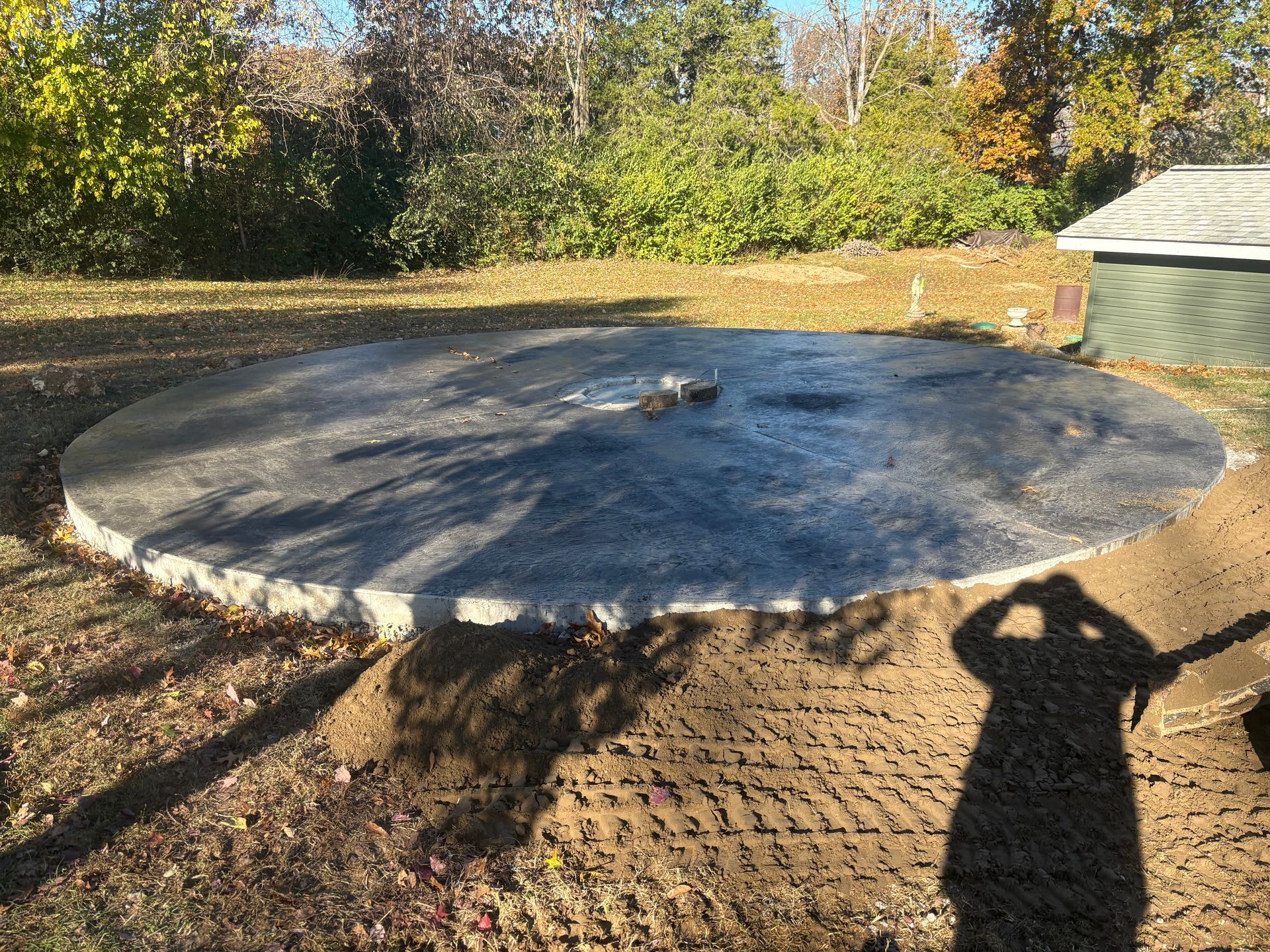 Large circular concrete slab with a small center opening, surrounded by dirt and trees in a sunny yard.