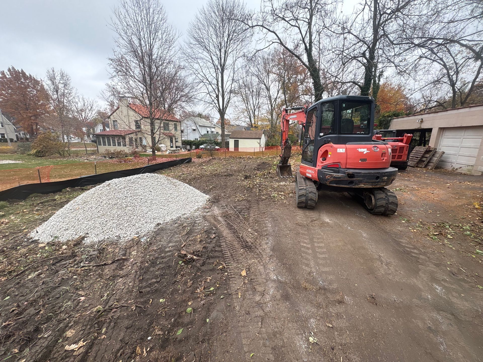 Red excavator on a dirt lot beside a gravel pile, with houses and bare trees in the background.