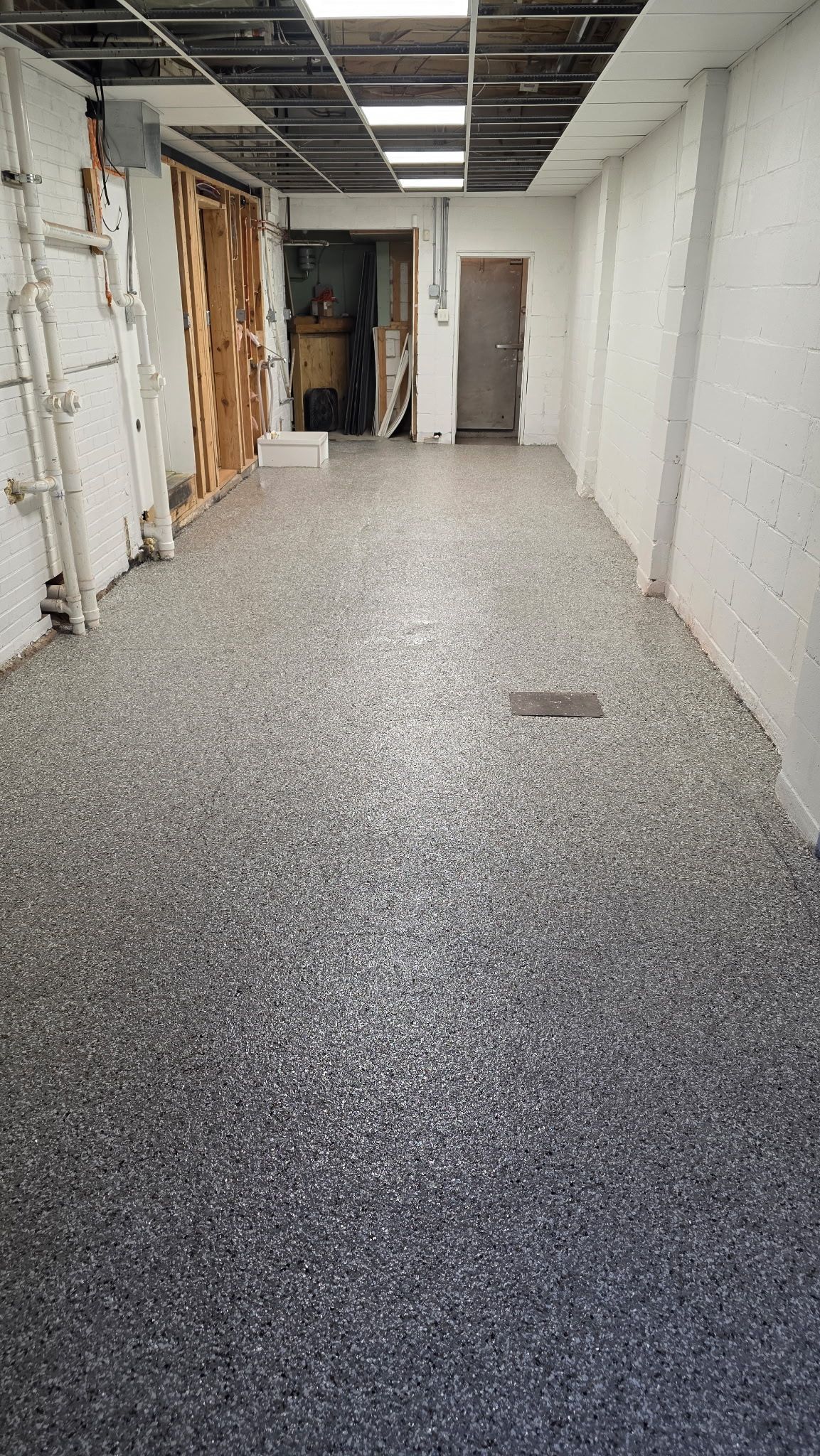 Narrow unfinished hallway with exposed walls, ceiling fixtures, and speckled floor.