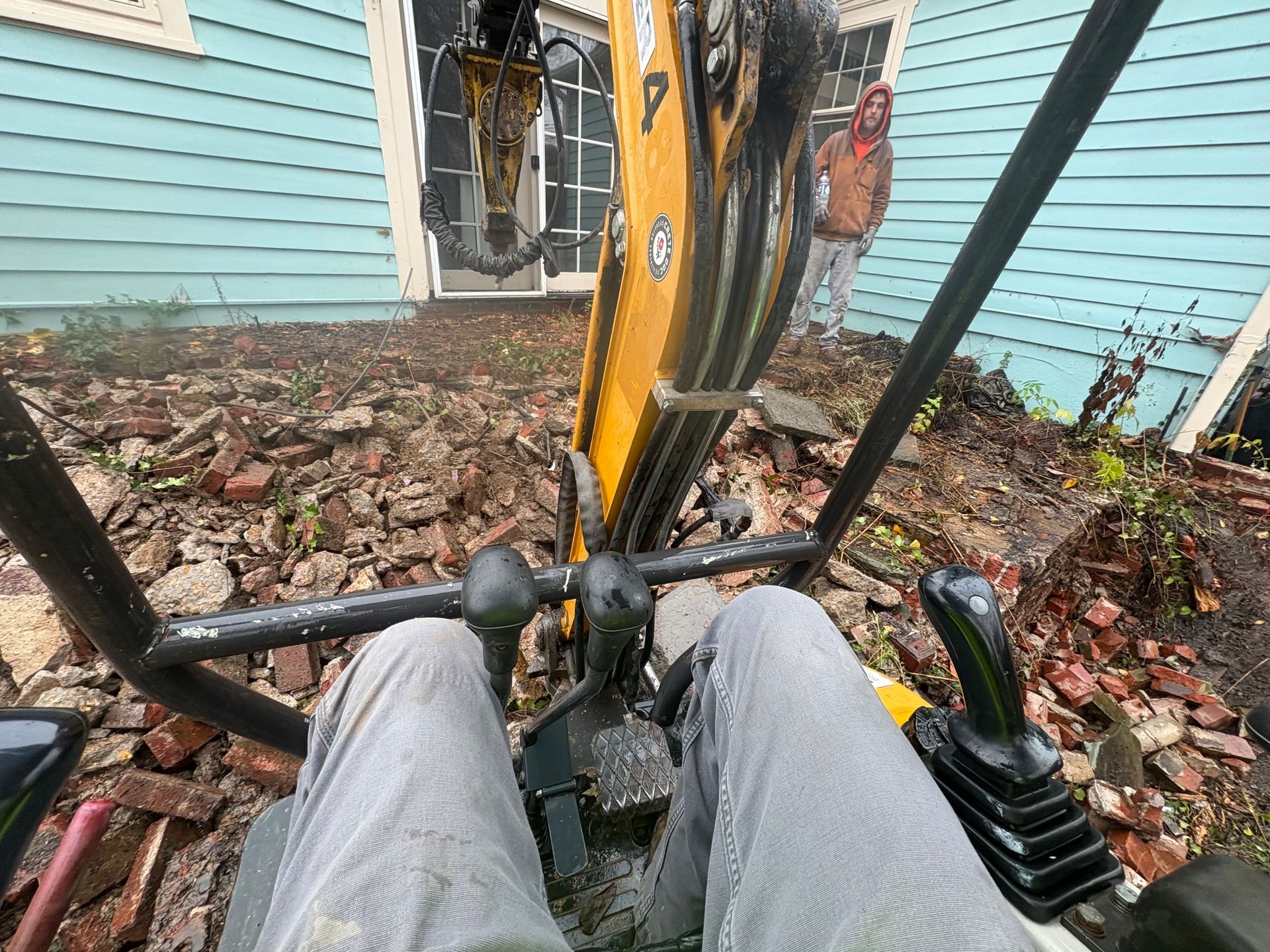 View from an excavator over muddy ground, with the operator’s legs, controls, and a blue house in front.
