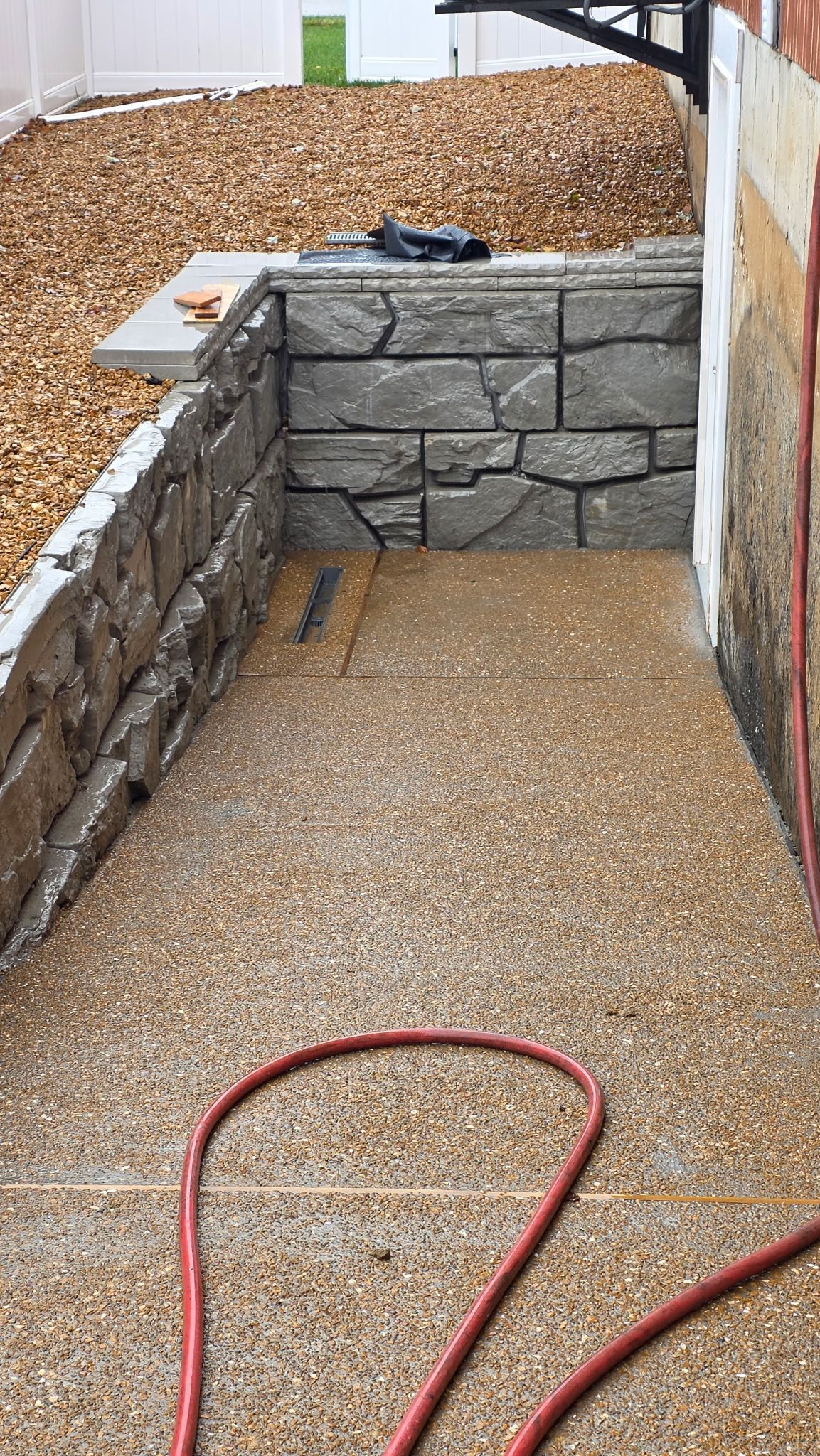 Narrow gravel path with low stone retaining walls and a red hose in the foreground.