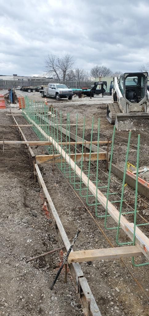 Construction site with wooden forms and rebar trench beside parked trucks under a cloudy sky.