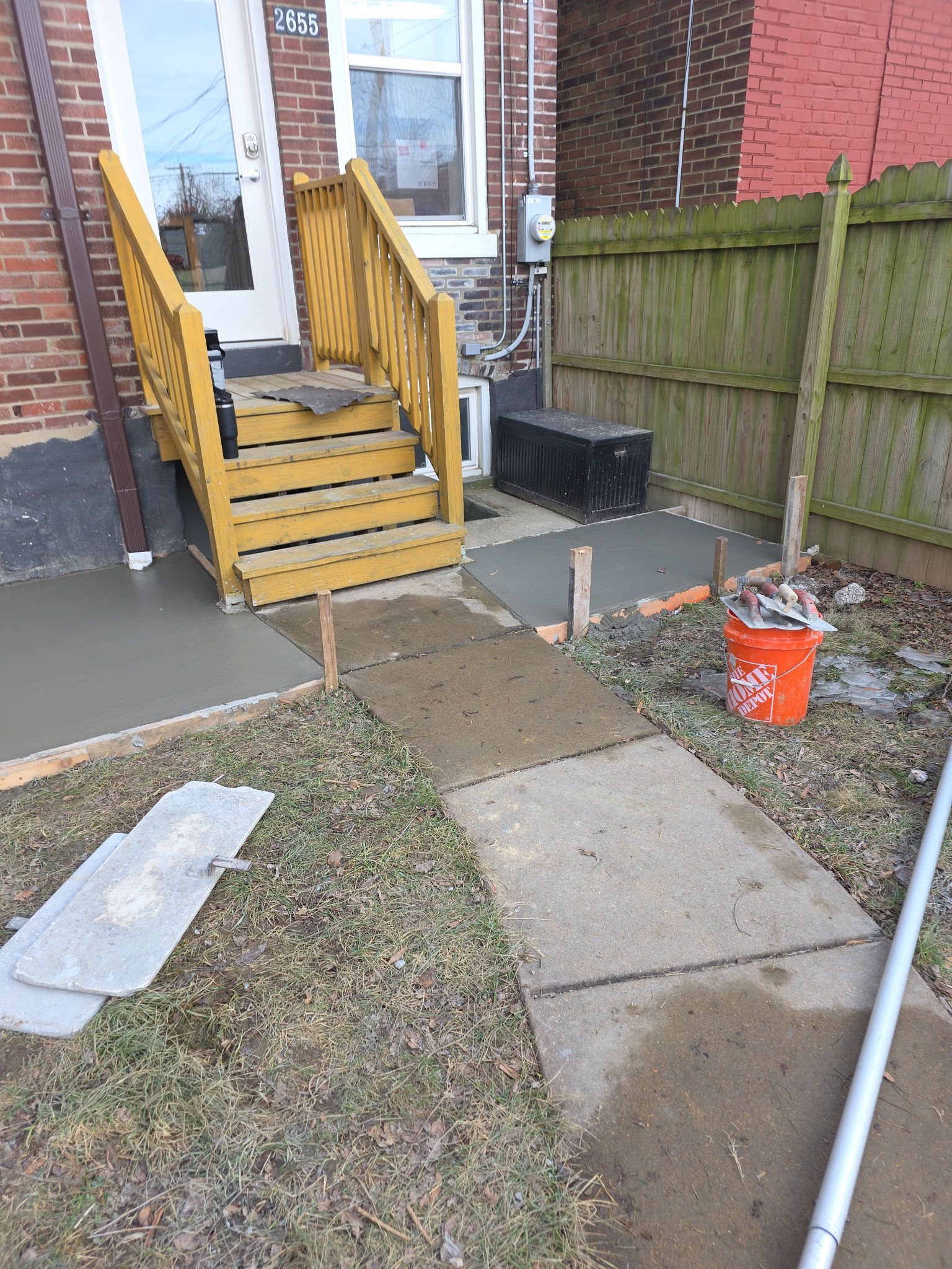 Front walkway to a house with yellow steps, concrete path, grass, and a fenced side yard with an orange bucket.