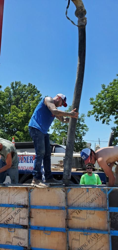 Worker in blue shirt pours concrete from a pump into a foundation wall under a clear sky.