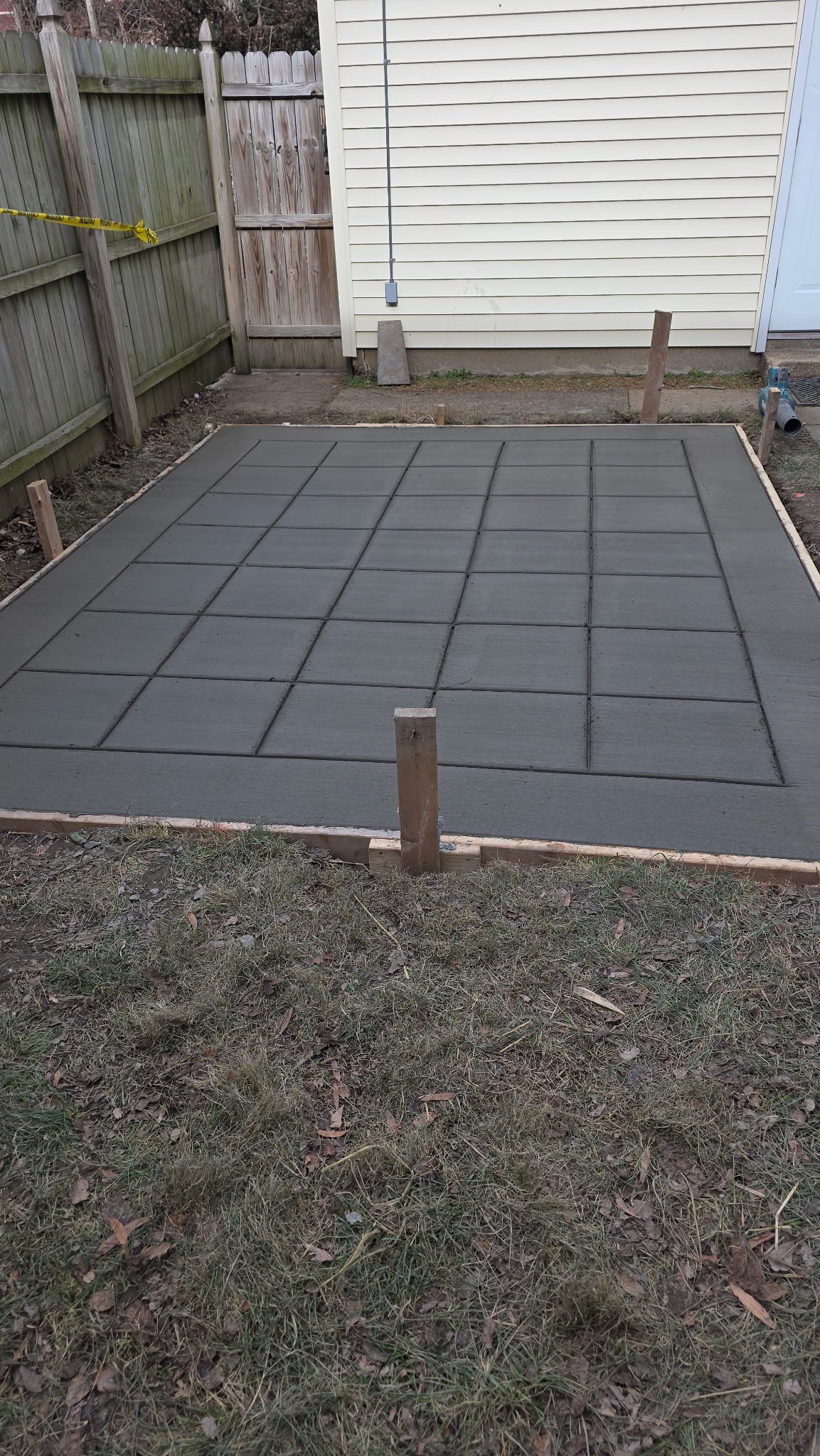 Small backyard patio with gray pavers, bordered by a fence and shed, and a wooden stake in the center.