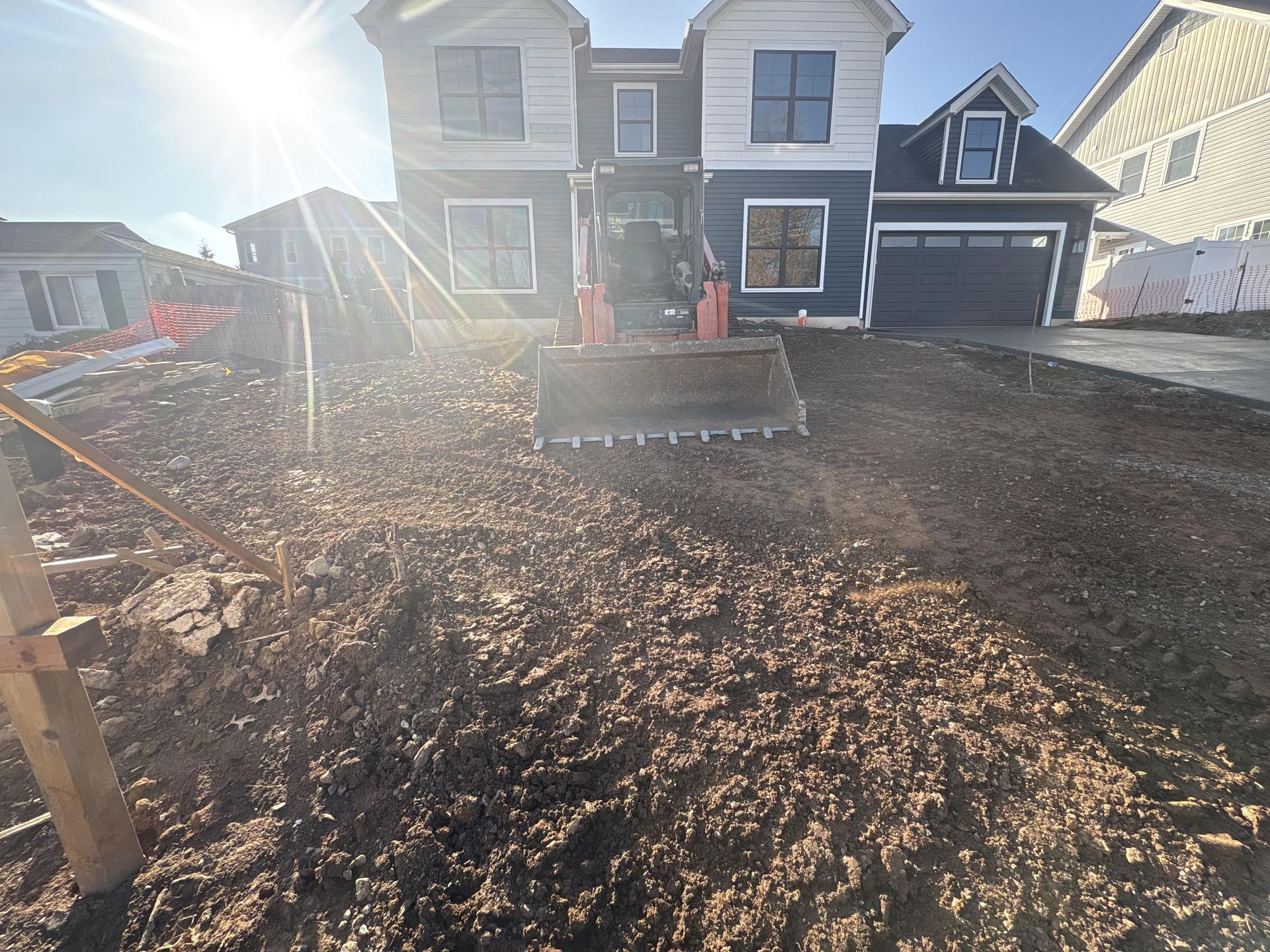 Front view of a two-story house with a muddy front yard and construction equipment in bright sunlight.