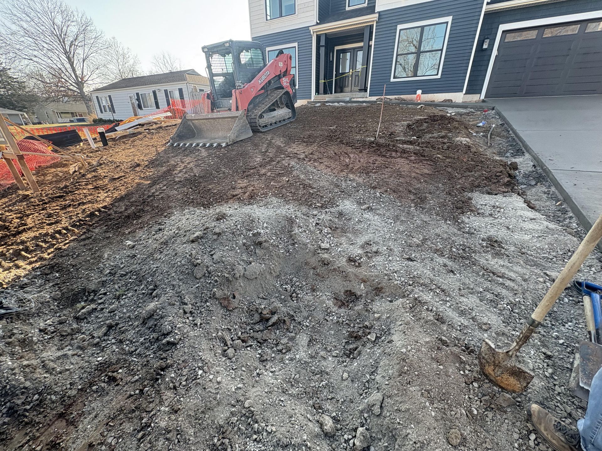 Construction site with excavator leveling dirt beside a house and driveway.