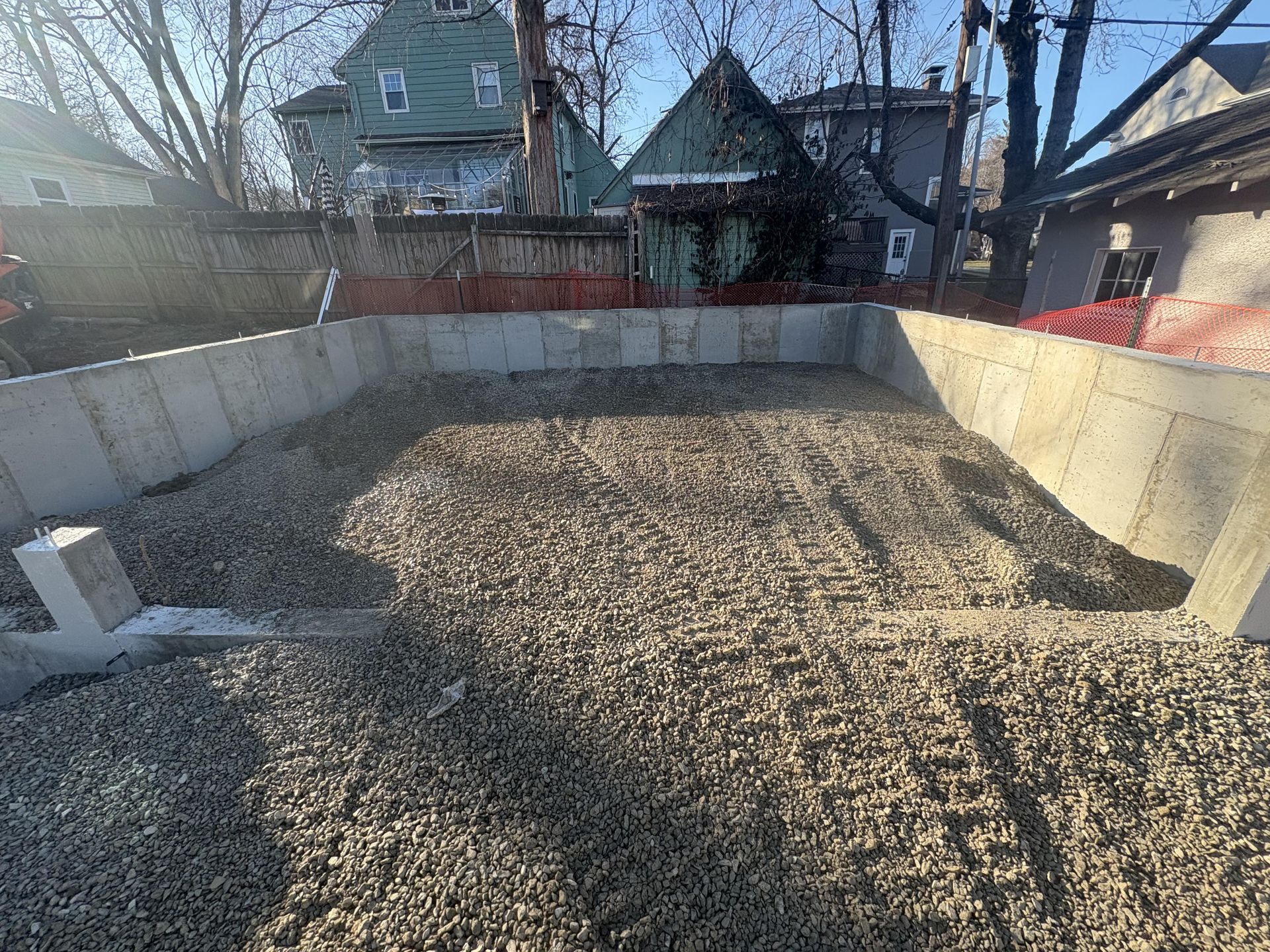Empty gravel-lined backyard pool under construction, with concrete walls and houses in the background.