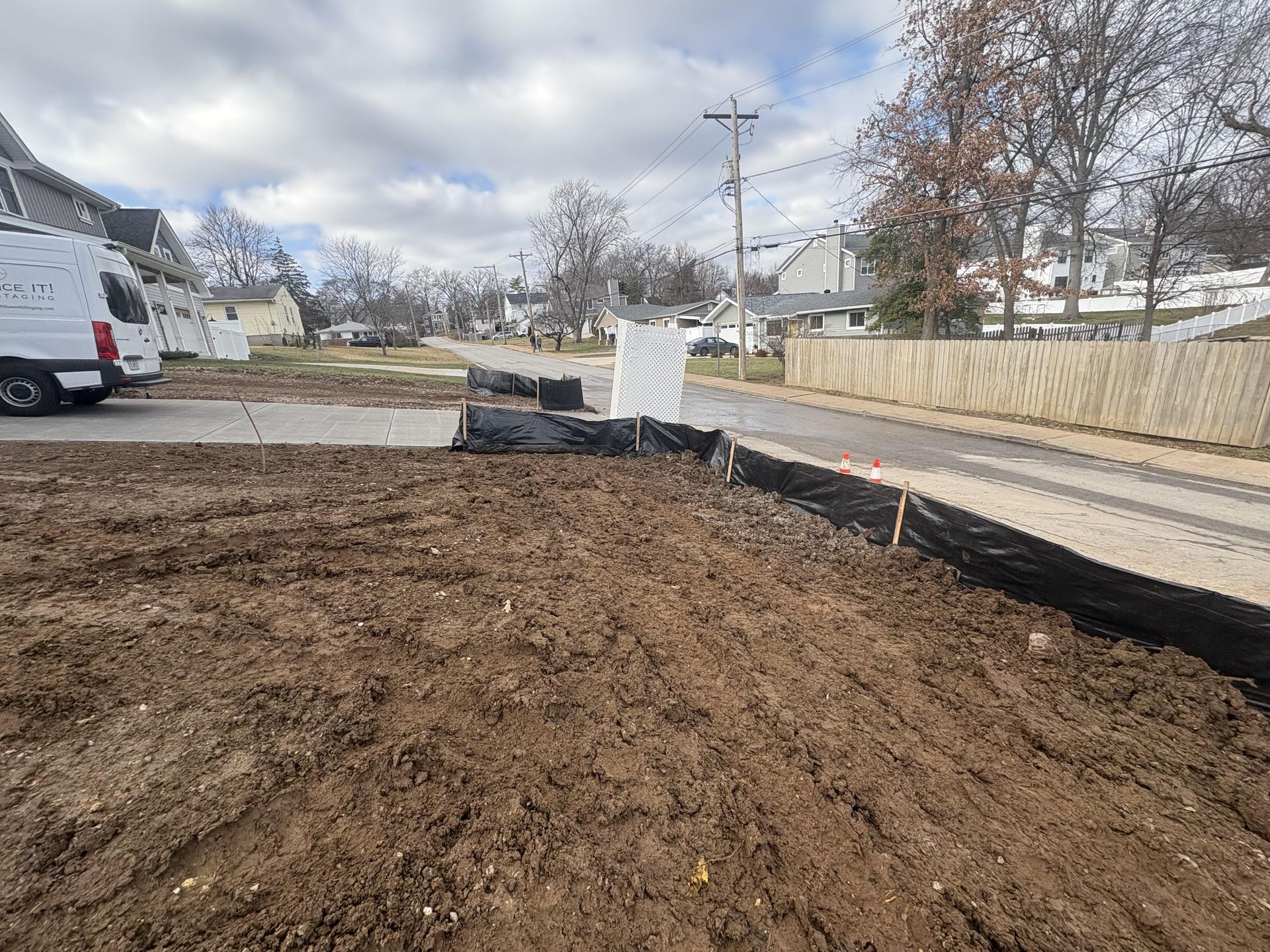 Newly graded dirt lot beside a street with a white van, fencing, and black erosion control barriers.