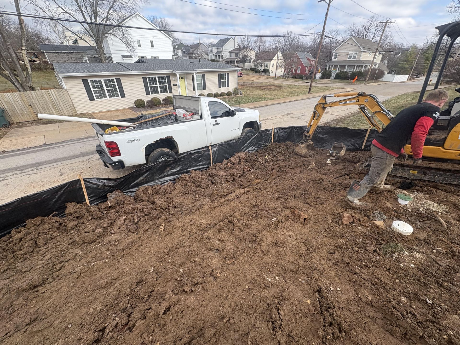 White pickup and excavator at a muddy construction site beside a house, with a worker shoveling soil.