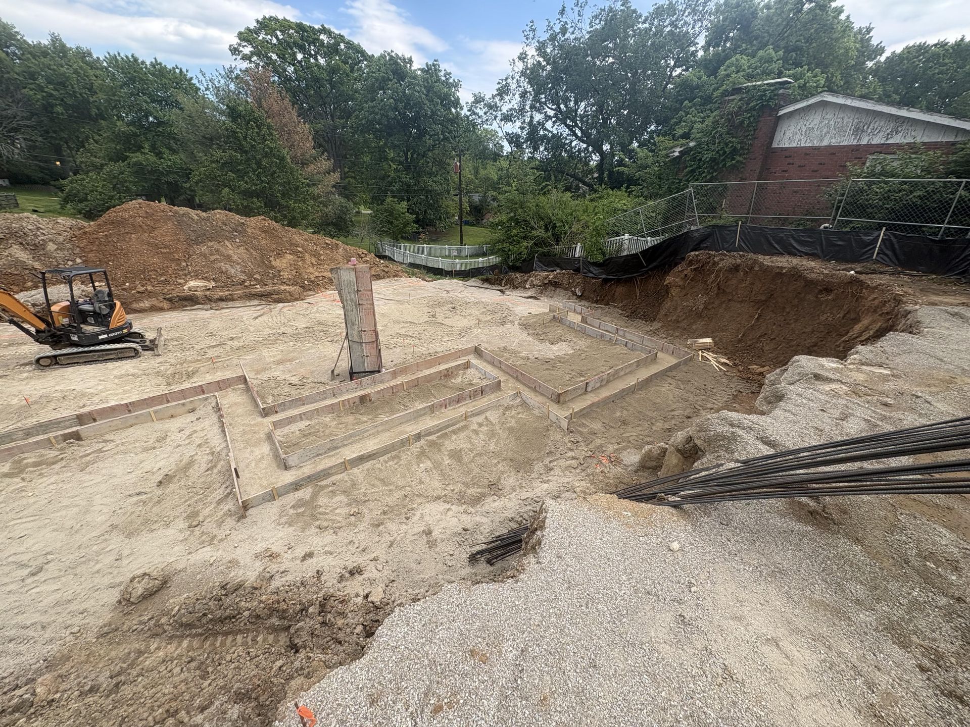 Excavation site with concrete foundation forms, dirt piles, and construction equipment under a cloudy sky.
