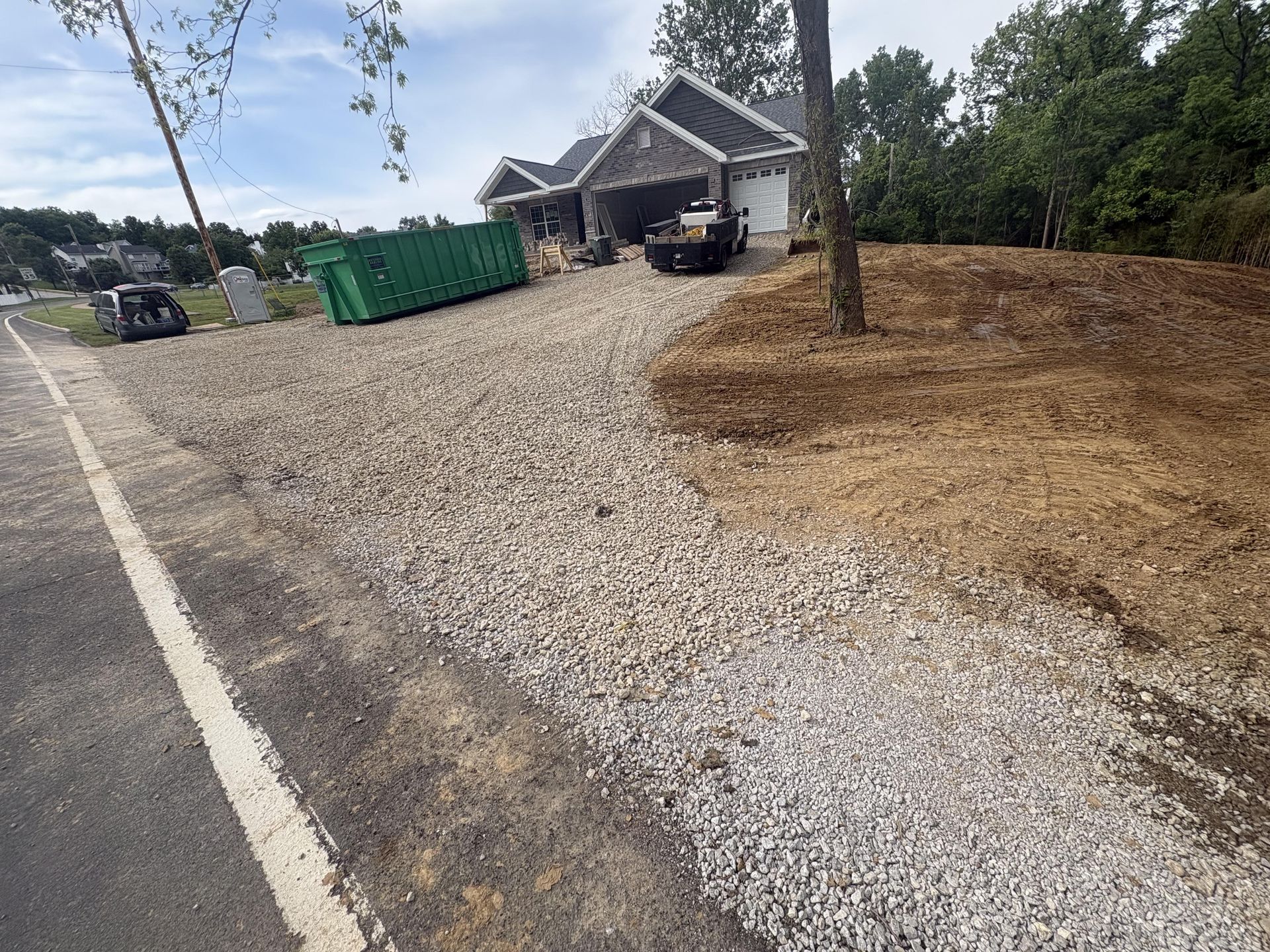 Gravel driveway leading to a house with a dumpster, parked vehicle, and a tree in a dry yard.