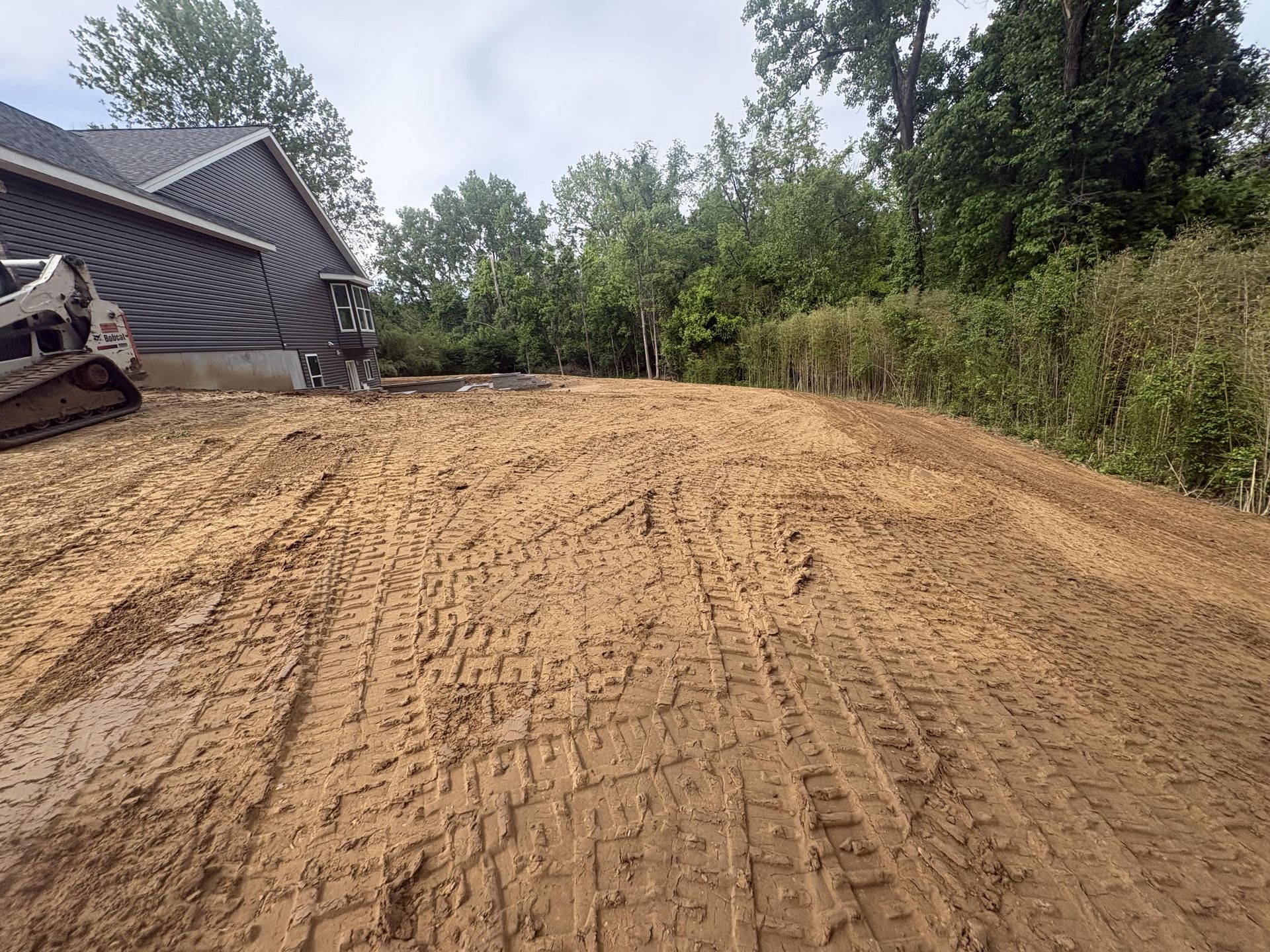 Freshly graded dirt lot beside a house, with trees and shrubs along the edge.