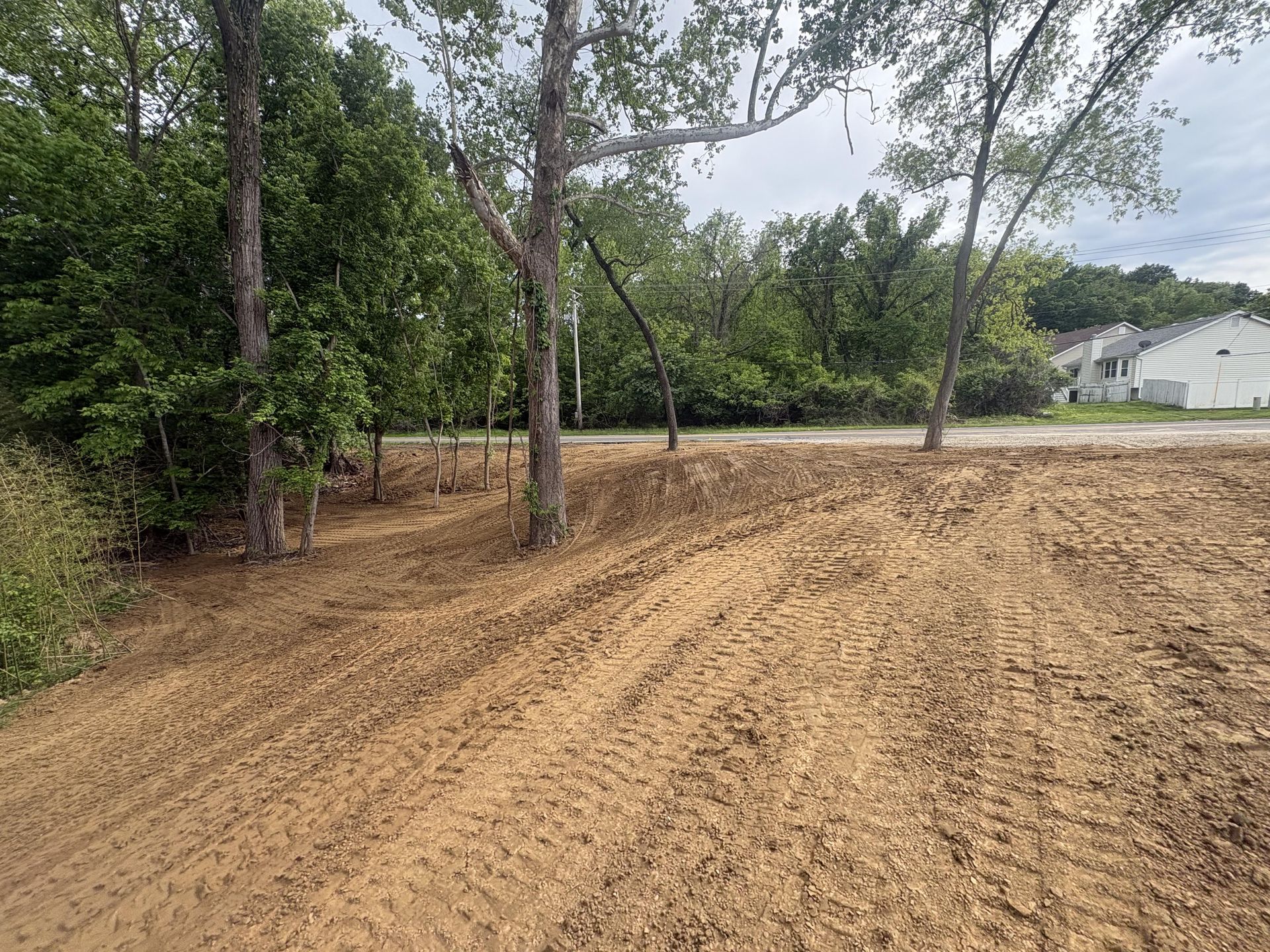 Cleared dirt lot with tire tracks beside trees and a white house under a cloudy sky.