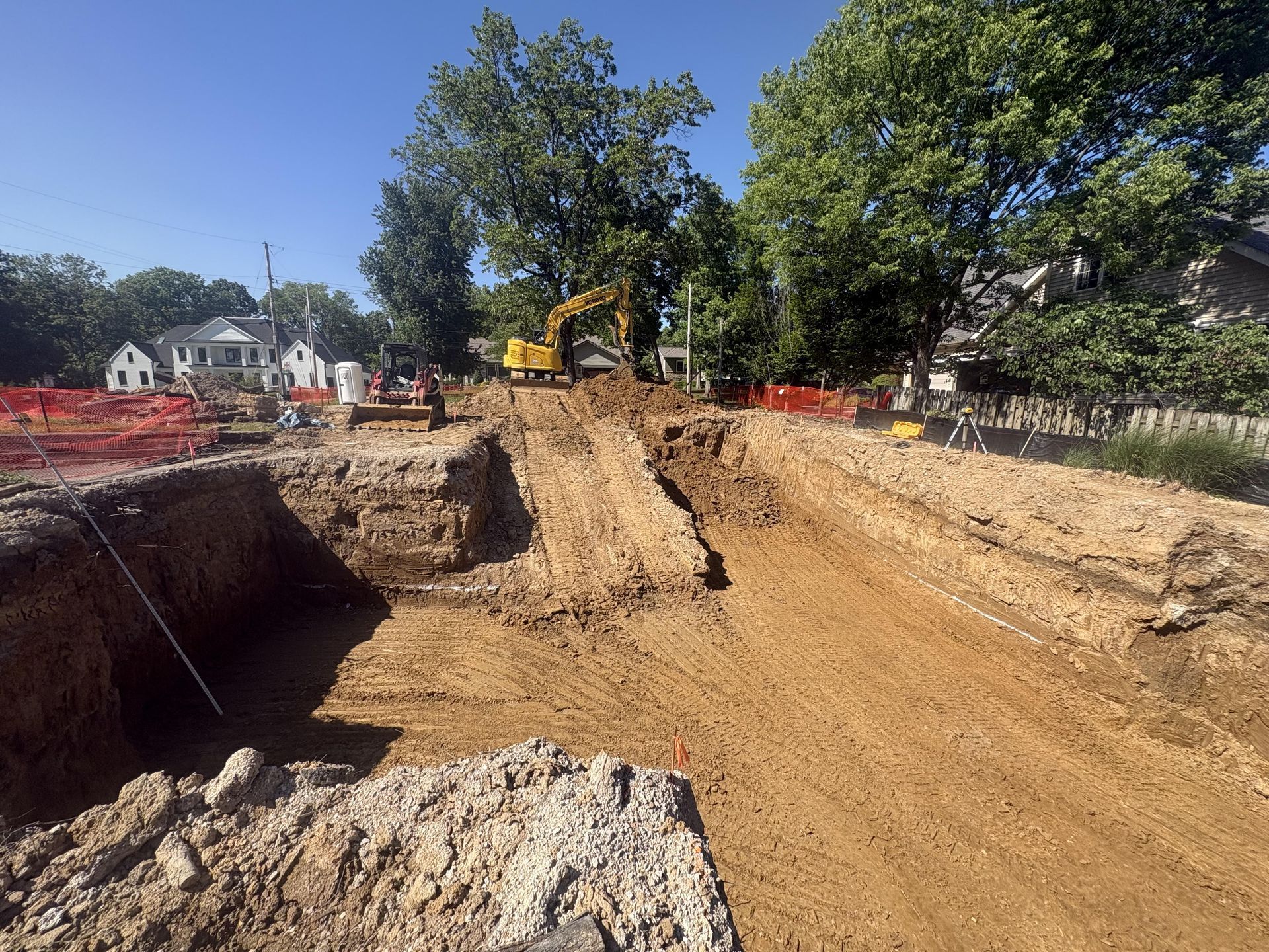 Construction site with excavator digging a deep trench beside a dirt road under a clear blue sky.