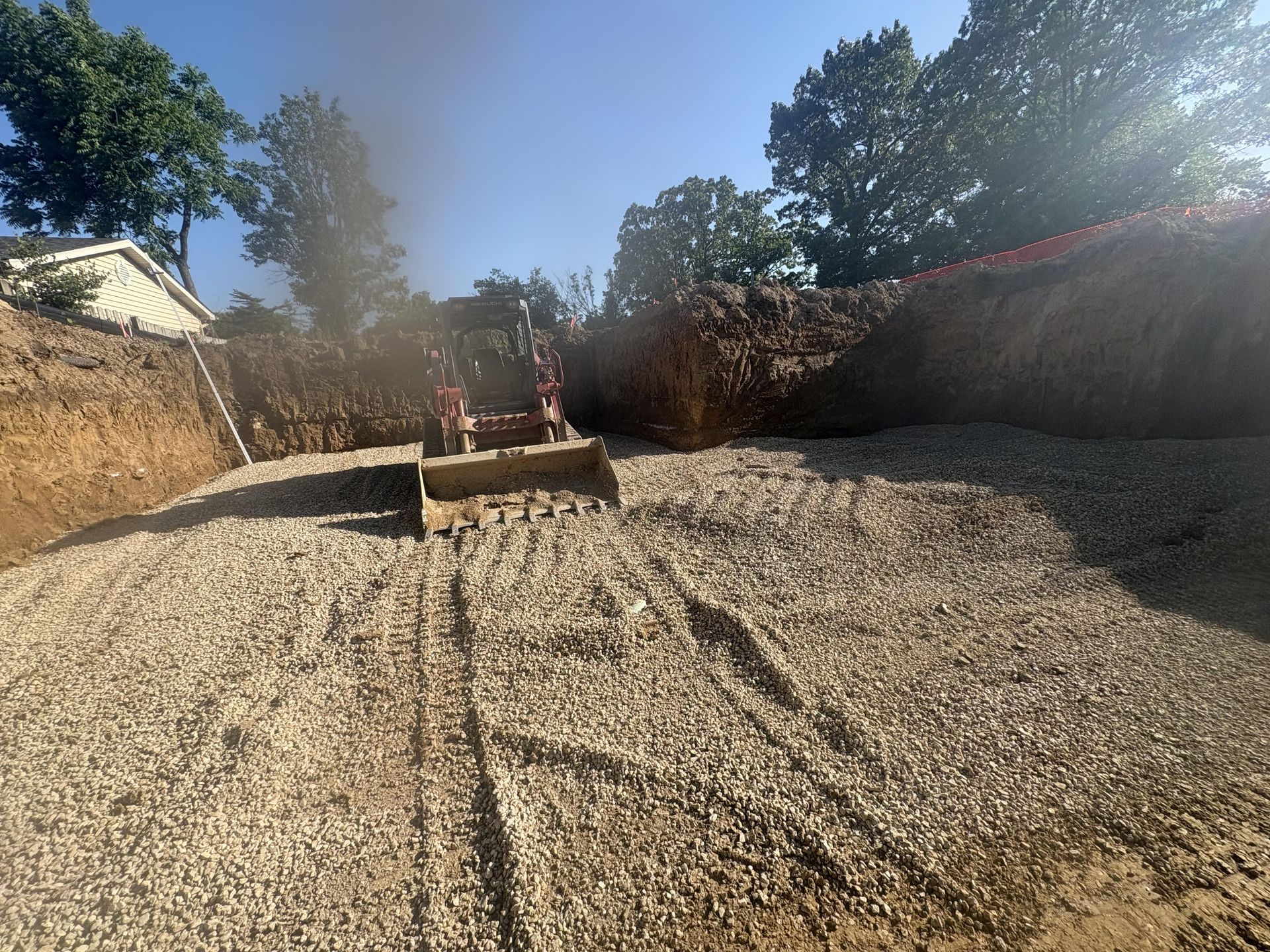 Construction site with an excavator spreading gravel across a sloped dirt area under a bright sky.