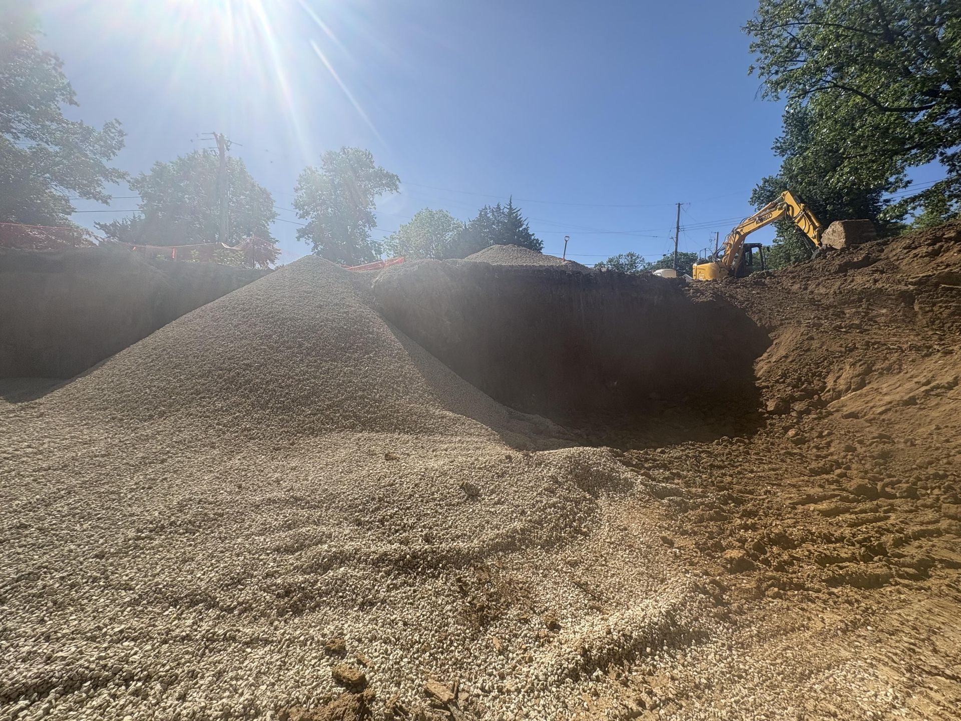 Wood chip piles in a bright outdoor yard, with an excavator on the right under a blue sky.