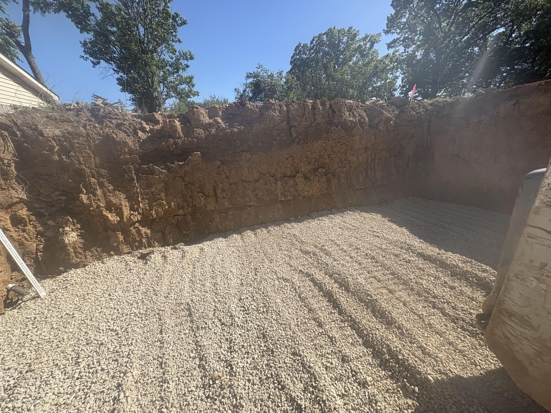 Fresh gravel spread in a dug-out construction area beside an excavator bucket and dirt retaining wall.