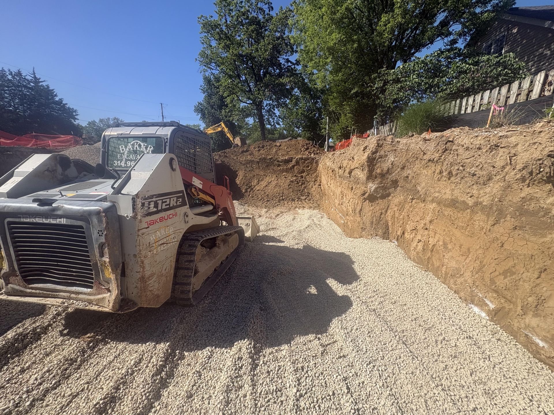 Construction loader spreading gravel in a narrow trench beside an earthen embankment under a blue sky.