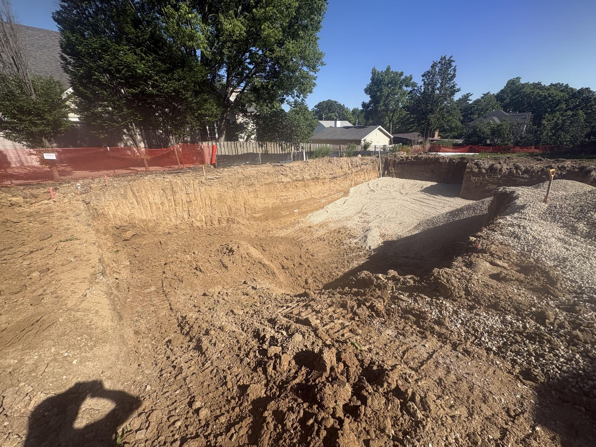 Dirt lot with a large excavated pit, trees and orange construction fencing in the background.