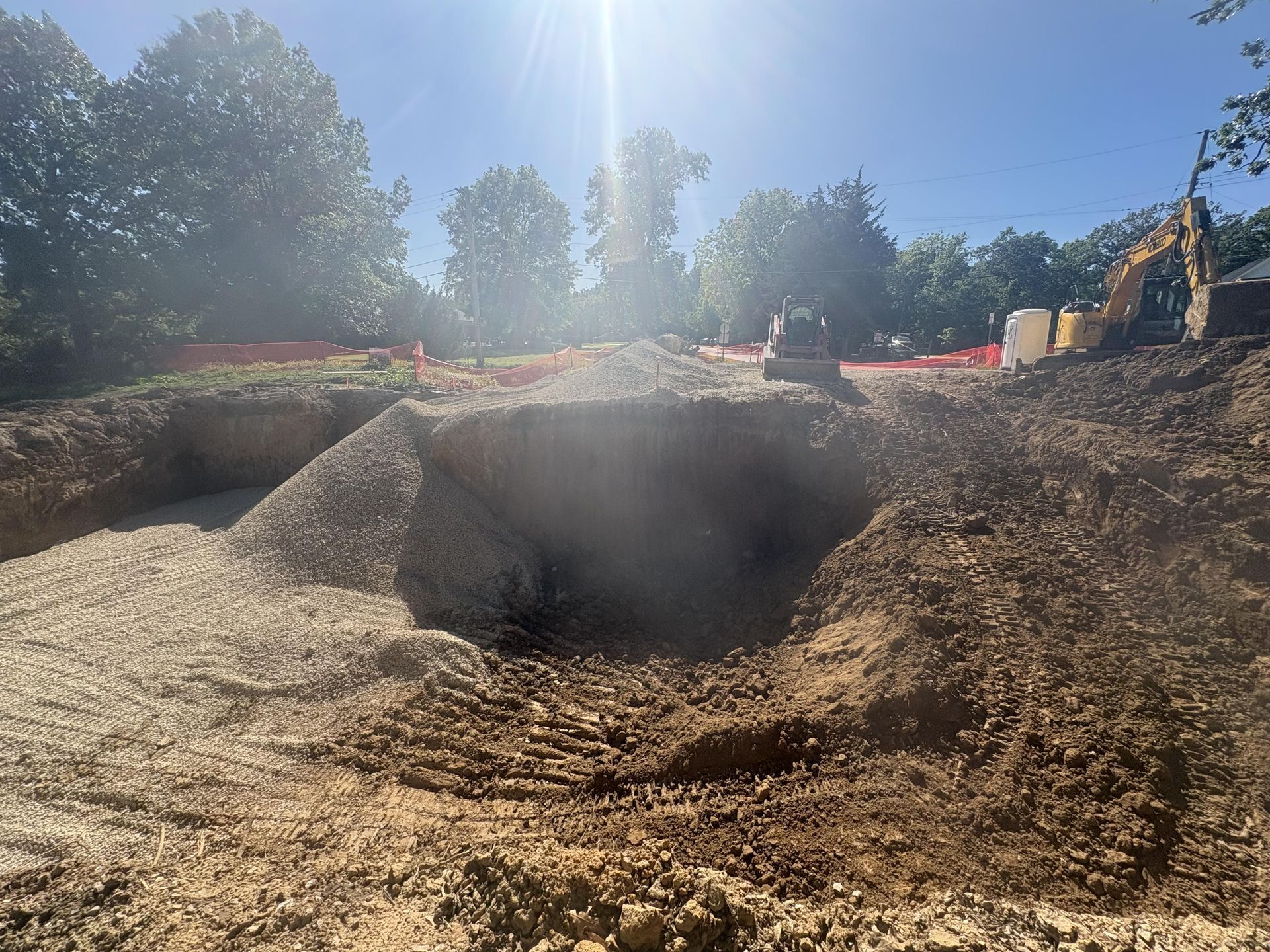 Large dirt excavation pit at a construction site under bright sun, with a backhoe in the distance.