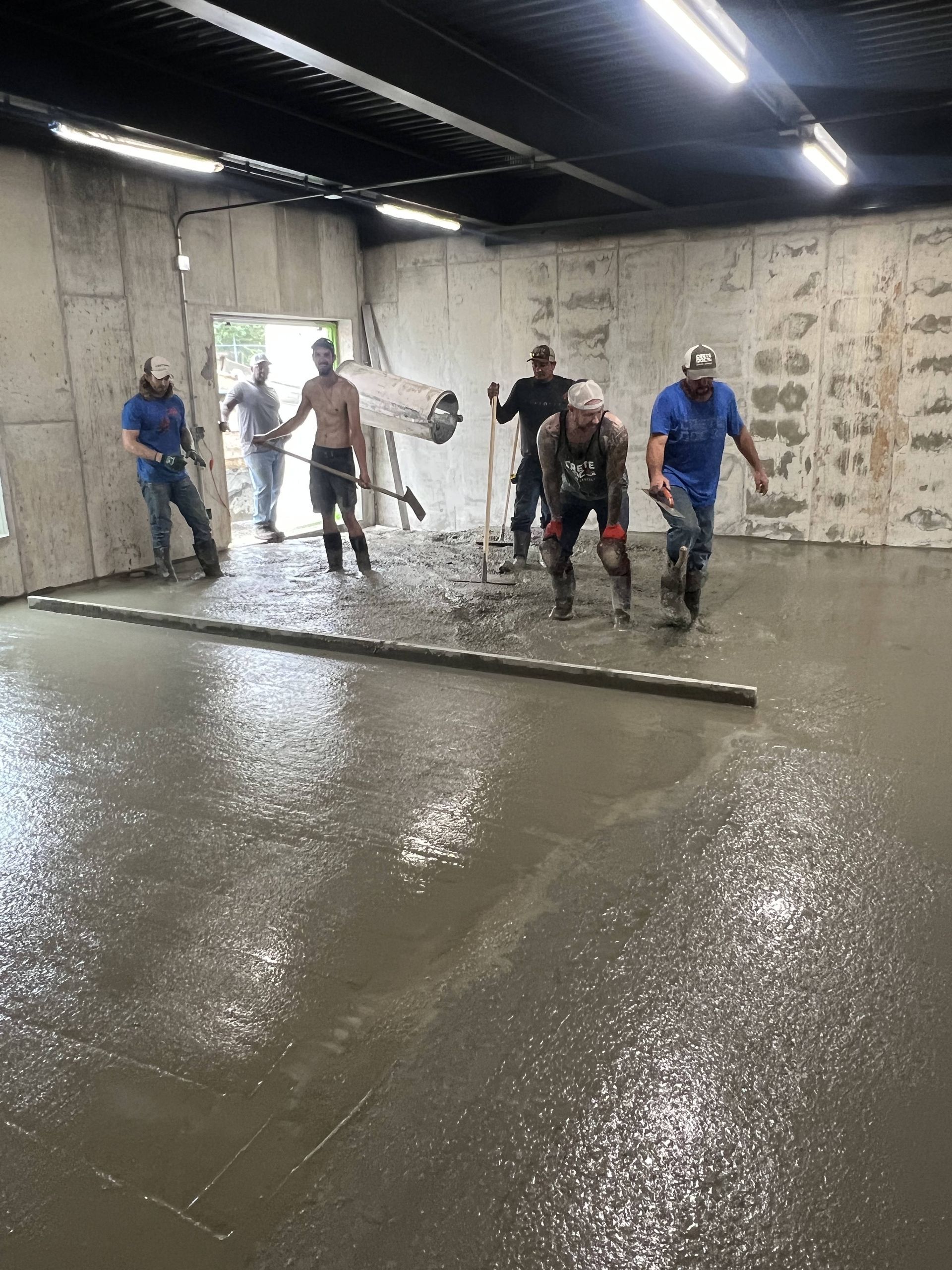 Workers spreading wet concrete in a large indoor room with walls covered in insulation.