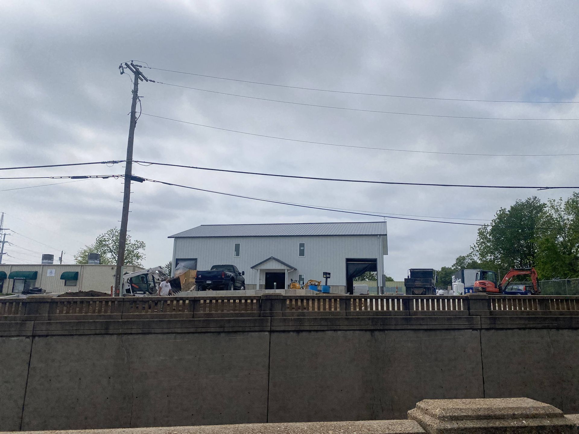 Cloudy roadside scene with a white building, utility poles, and parked trucks behind a concrete wall.