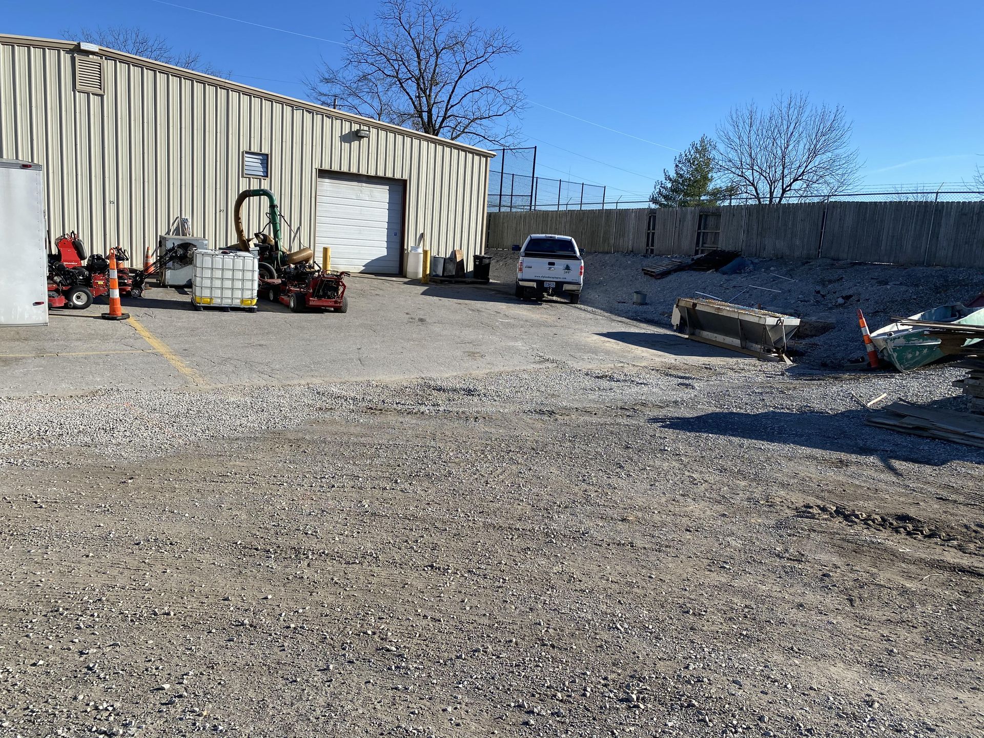Gravel lot with a white warehouse, parked vehicles, and construction equipment under a clear blue sky.