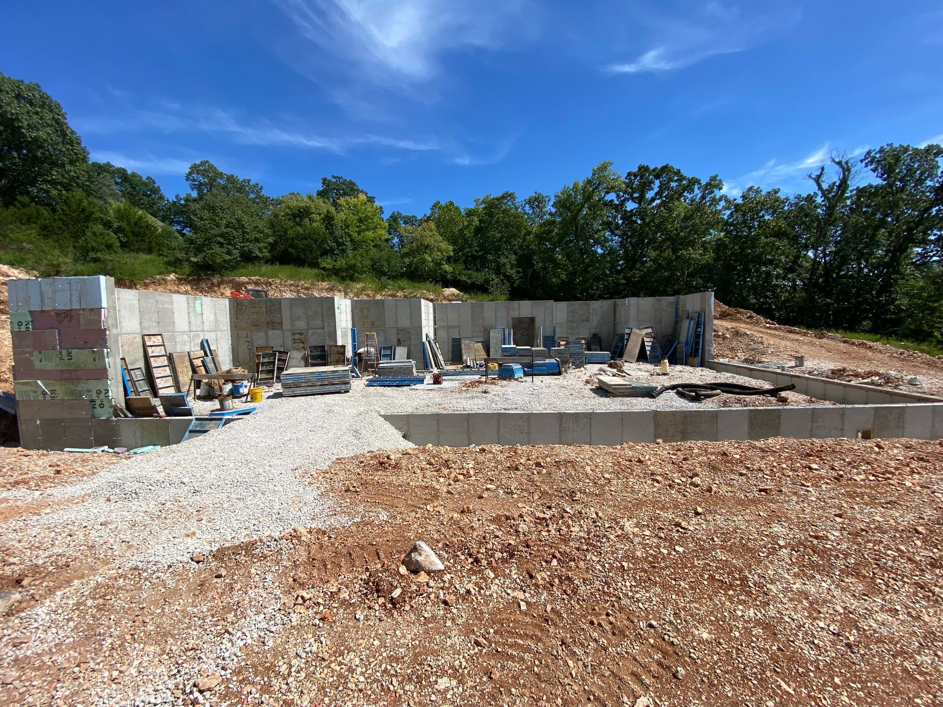 Outdoor construction site with concrete foundation, retaining wall, and gravel under a blue sky.