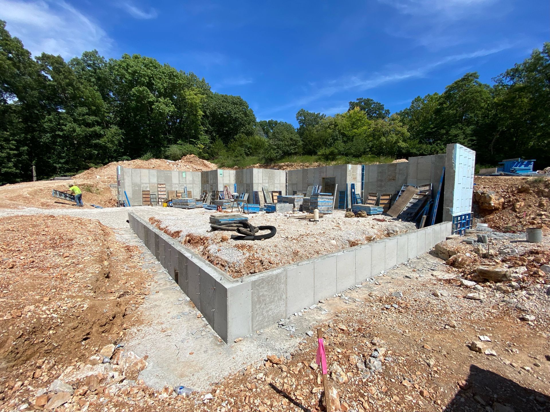 Foundation under construction with concrete walls, debris, and blue sky on a wooded site.