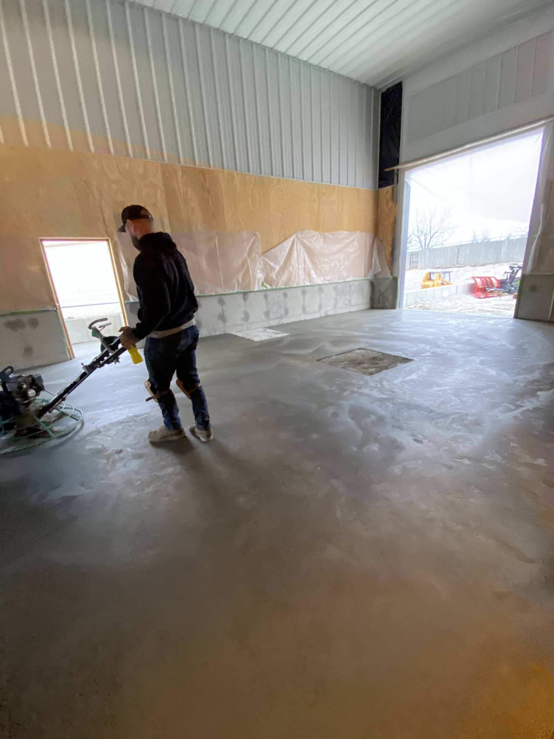 Worker polishing a concrete floor in a large empty warehouse with open doorway.