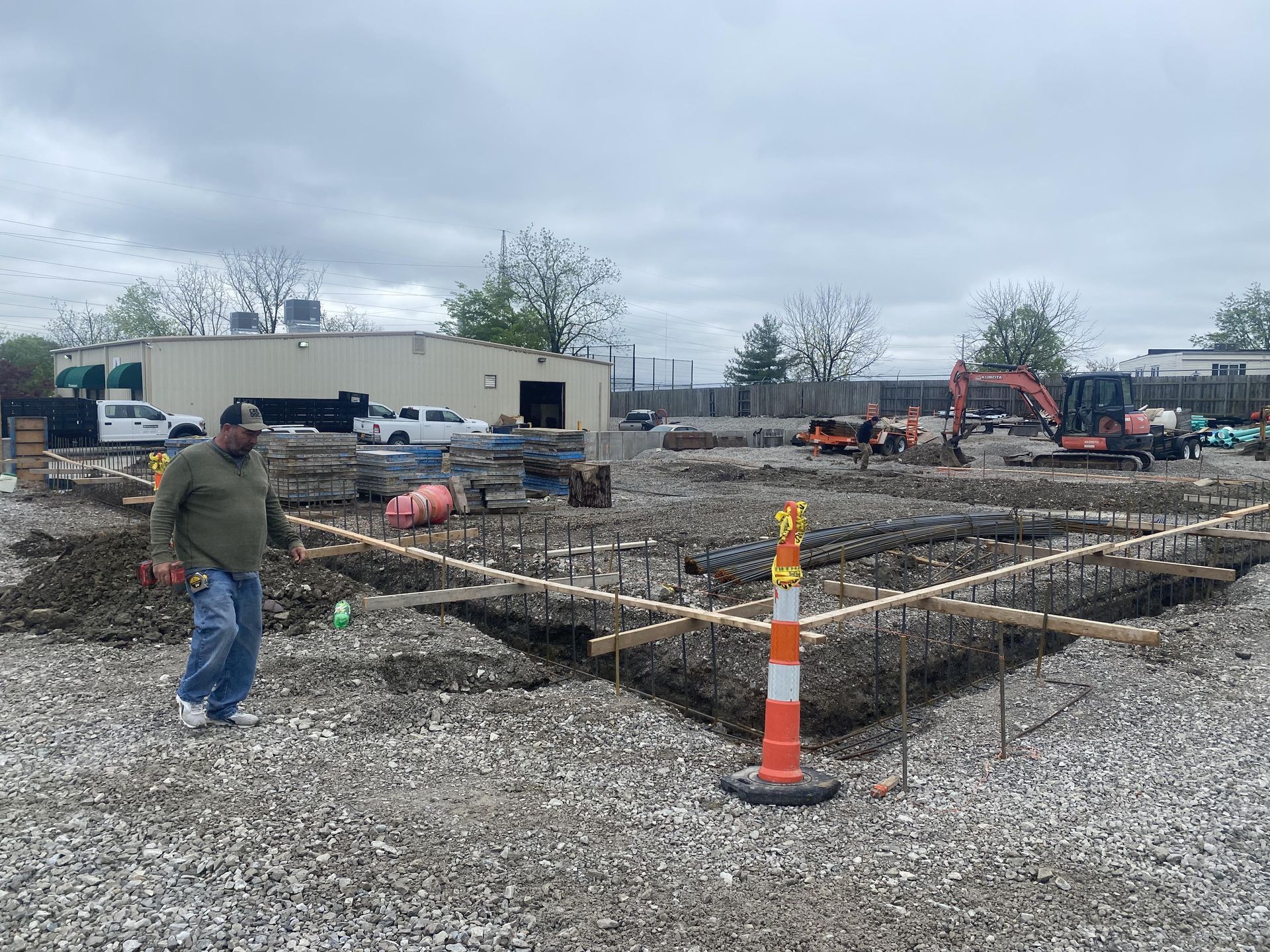 Construction site with rebar foundations, gravel ground, and a worker in a hard hat under cloudy skies.