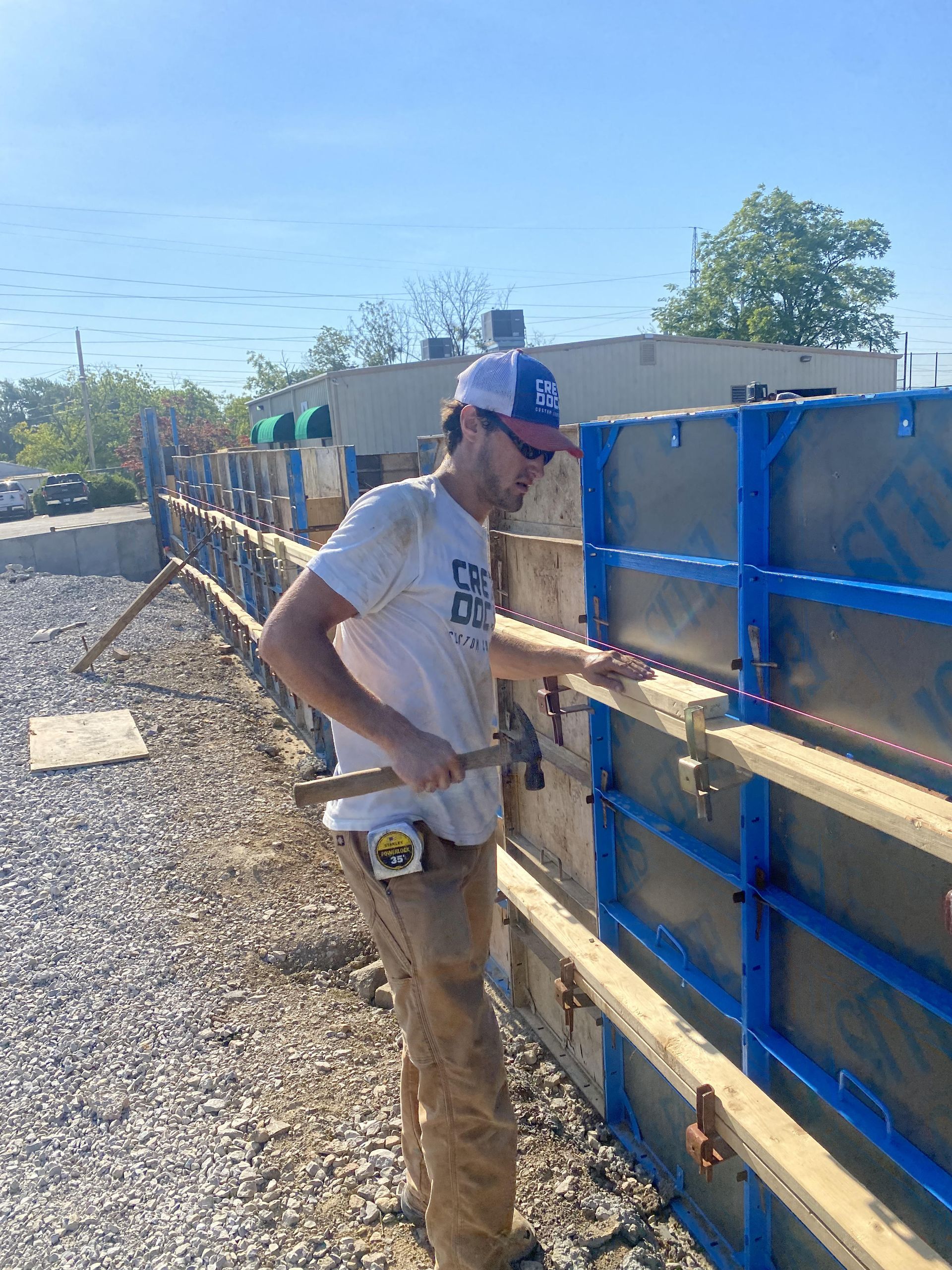 Worker in a hard hat measuring a wooden fence beside a gravel path under a clear blue sky.