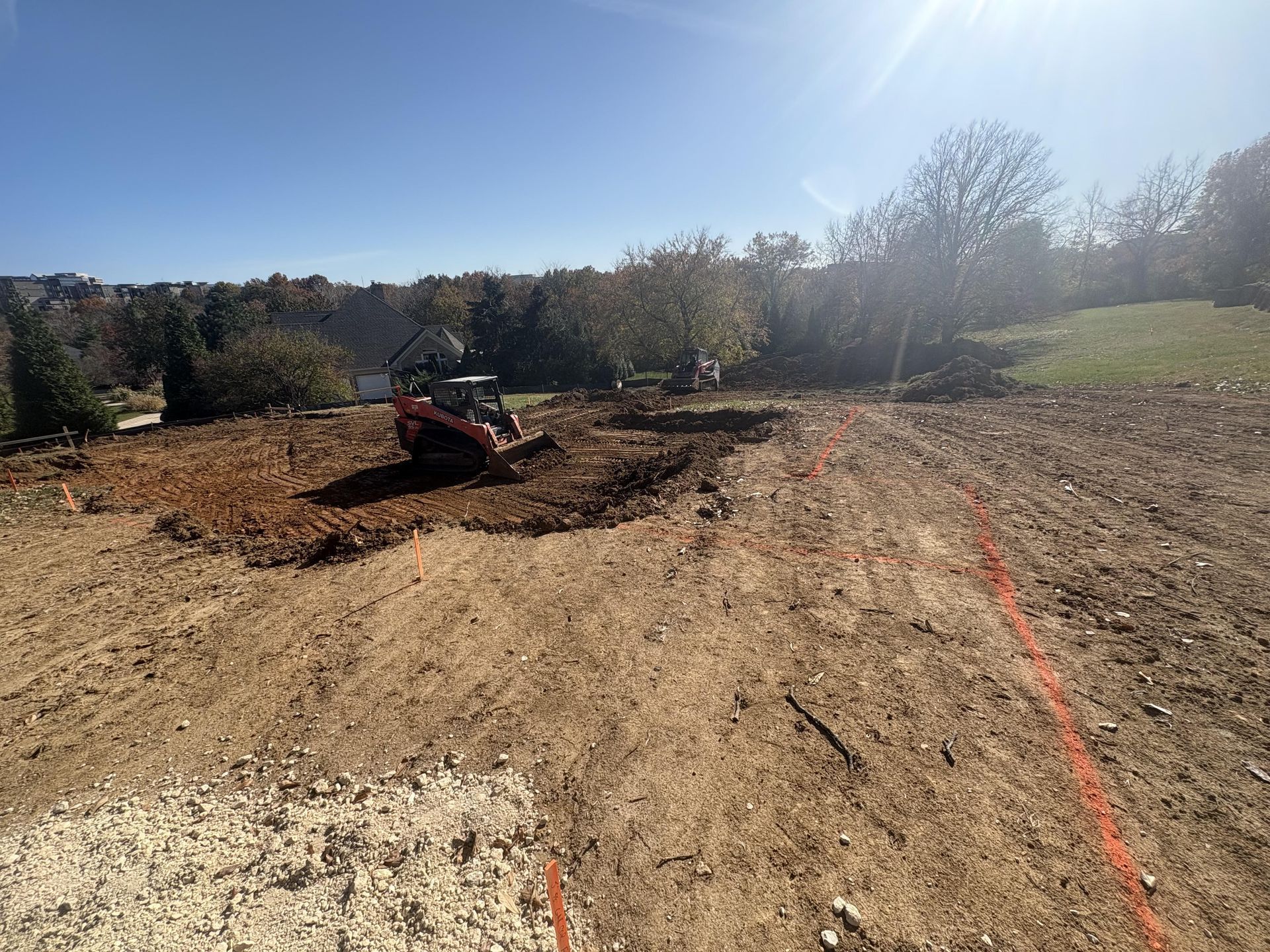 Excavator grading a dirt lot on a sunny hillside with trees in the background.