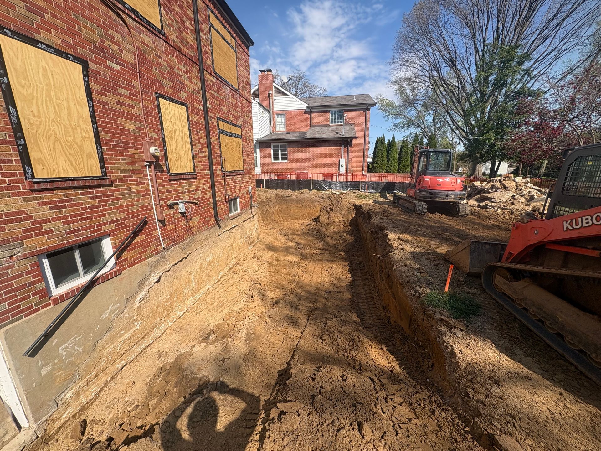 Backyard excavation beside a brick house, with a dirt trench and construction equipment.