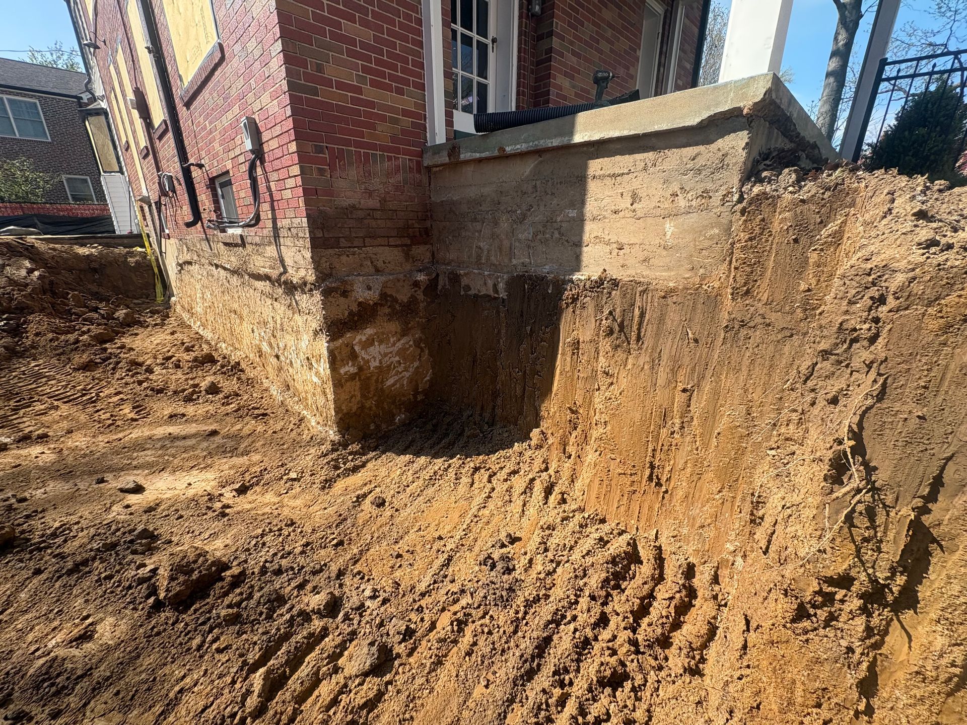 Excavated dirt foundation beside a brick house, with a dug-out trench and muddy ground.