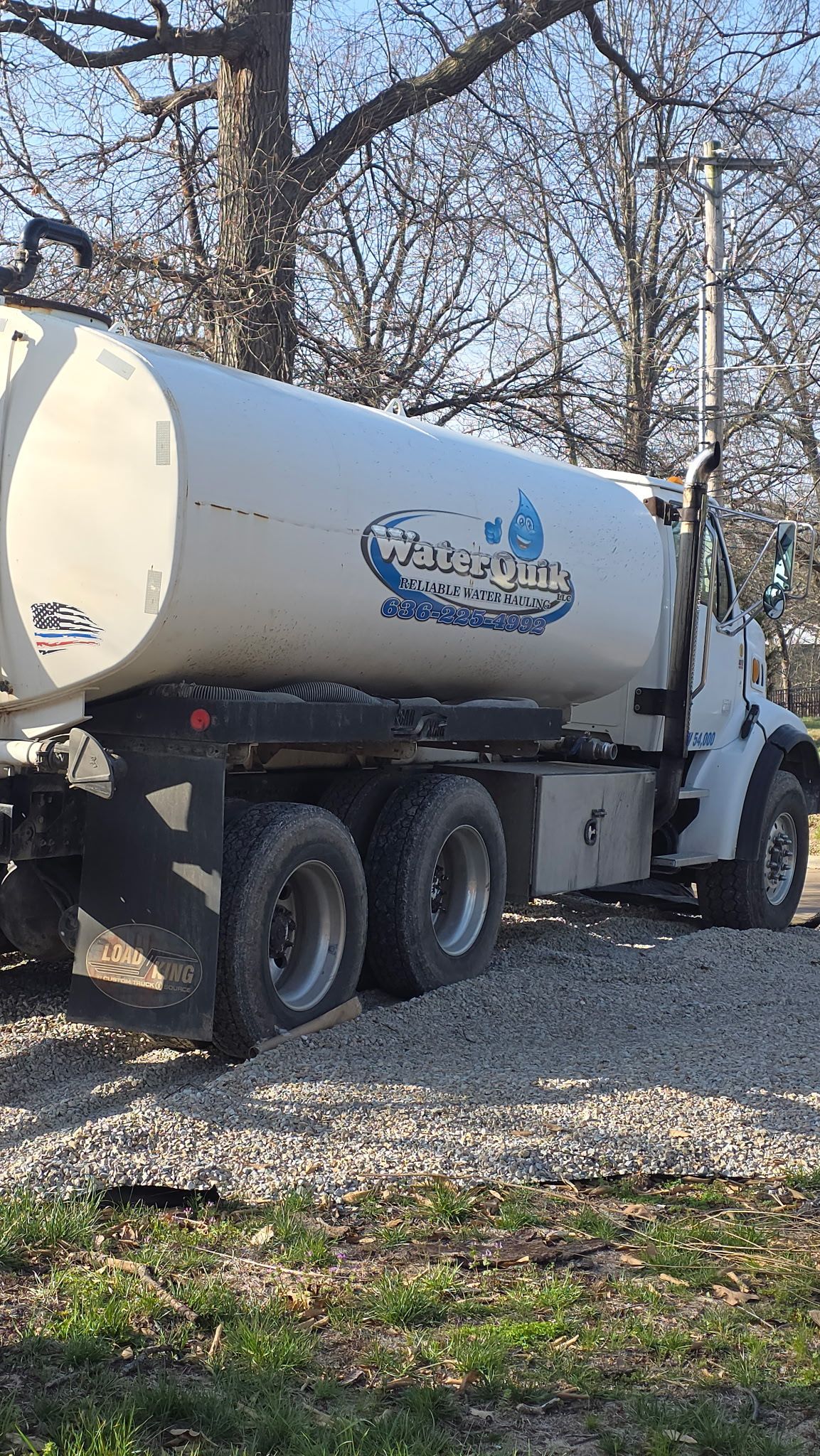 White vacuum truck parked on gravel in a wooded area, with bare trees in the background.
