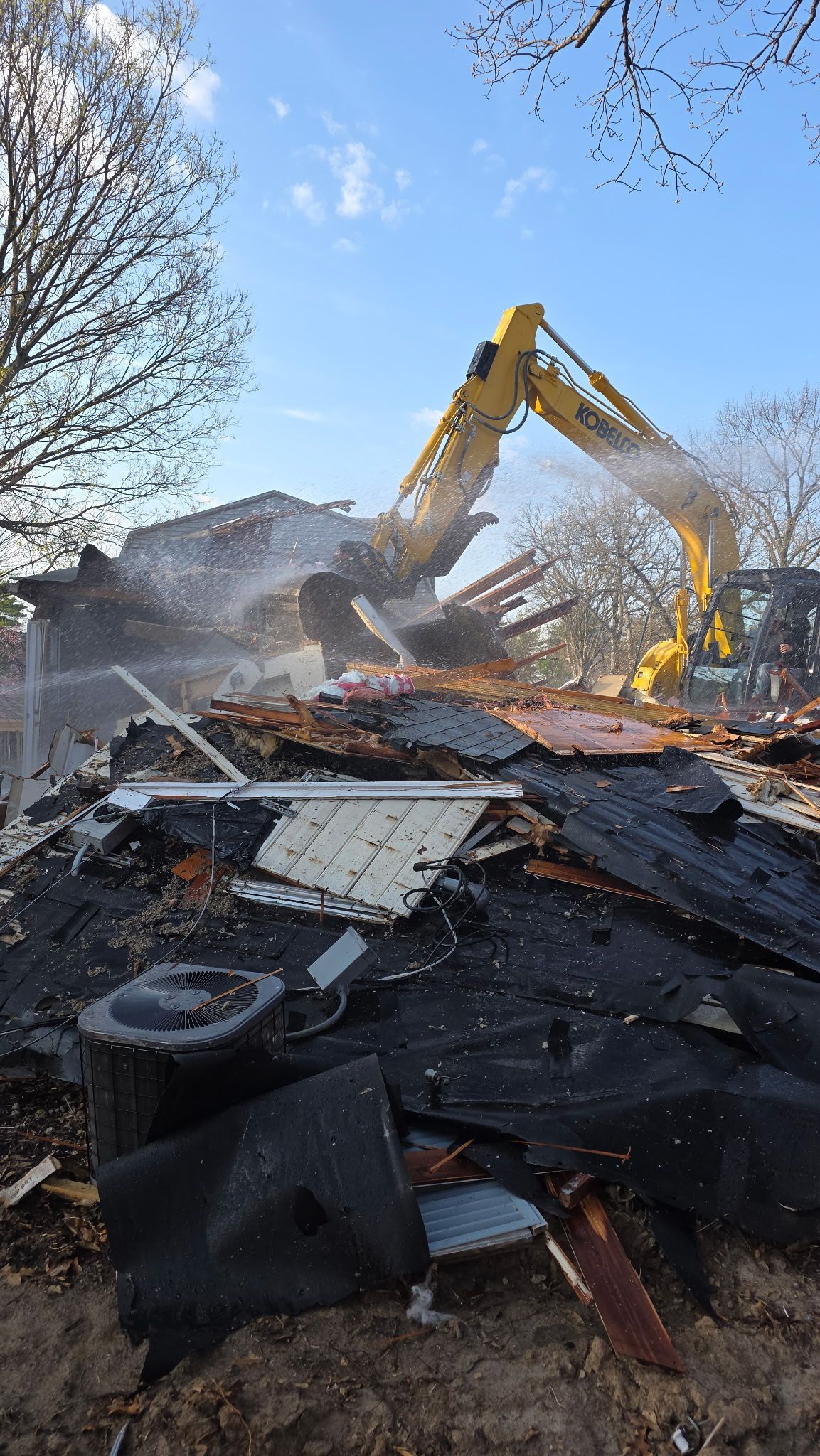Demolition site with excavator clearing debris amid smoke and rubble under a blue sky.