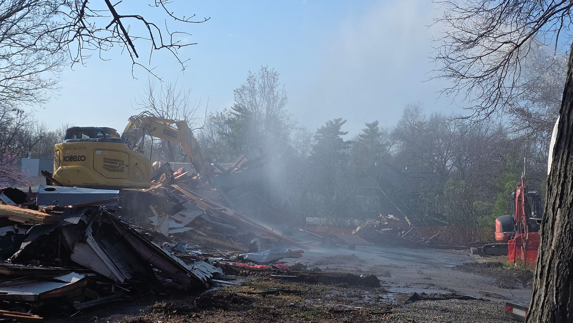 Demolition debris smolders on a roadside, with smoke rising beside bare trees and heavy equipment.