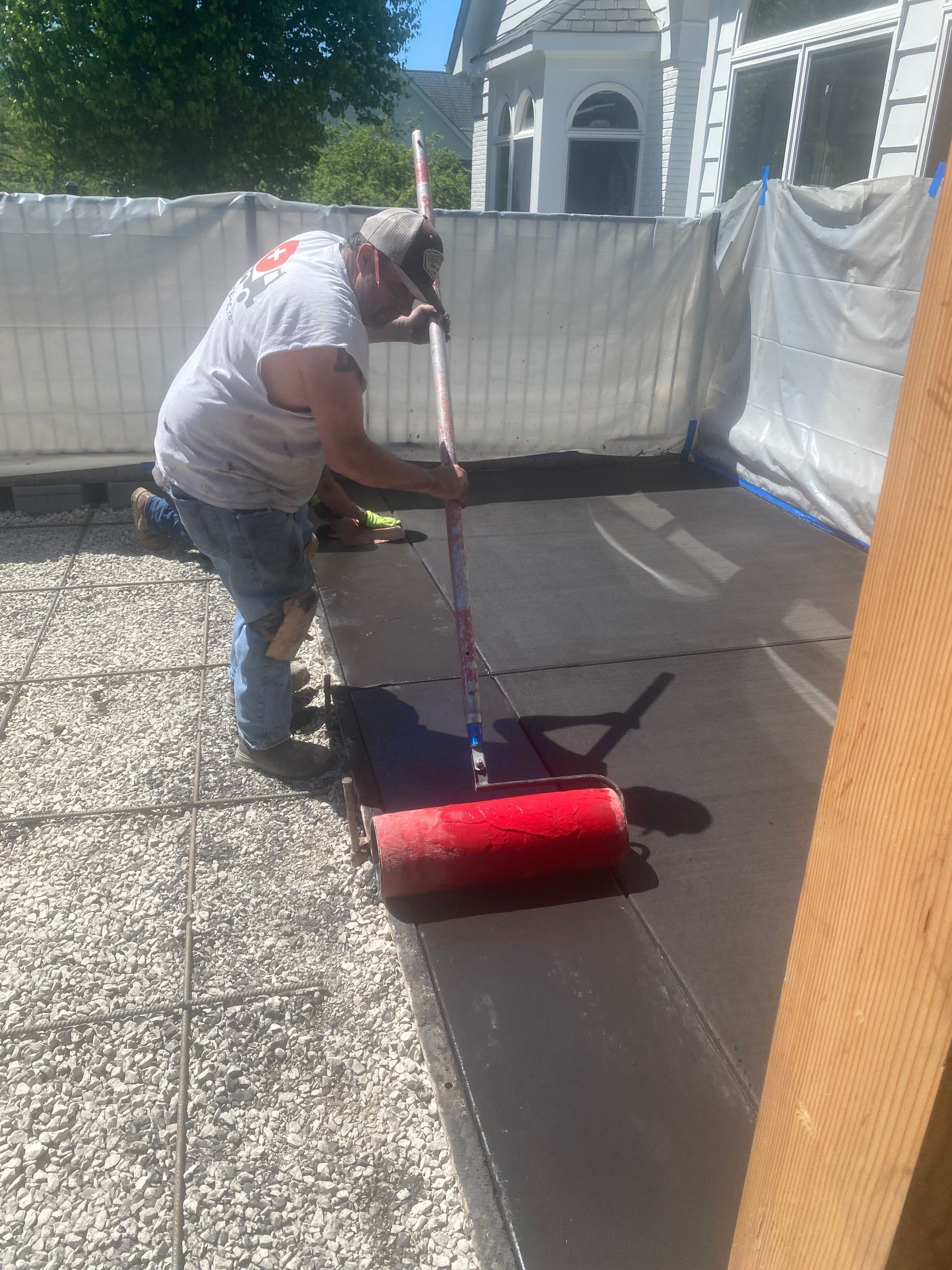 Person painting a dark driveway red with a roller outdoors beside a white fence.