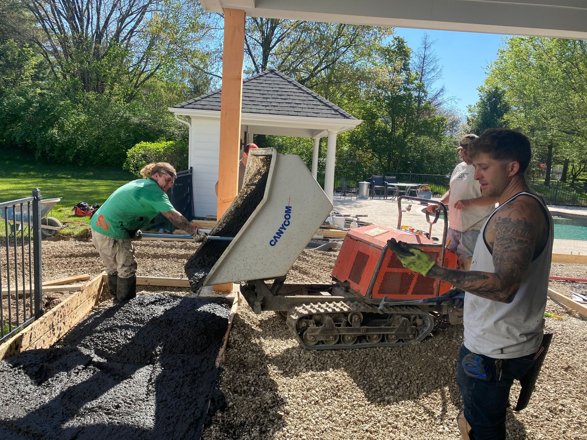 Two workers moving gravel with a wheelbarrow and mini excavator beside a gazebo outdoors.