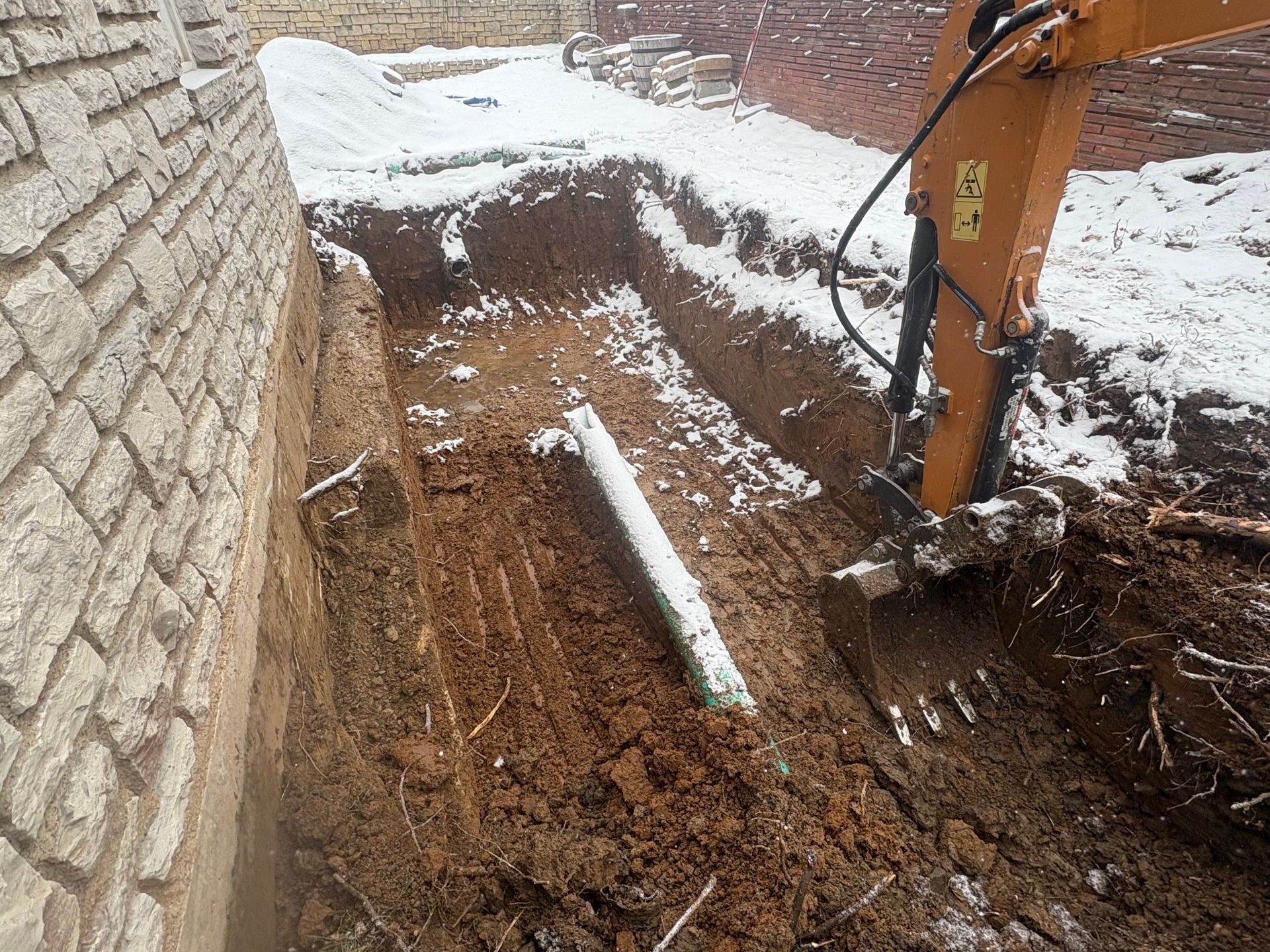 Excavator digging a snowy trench beside a stone wall, with exposed soil and a pipe in the ground.