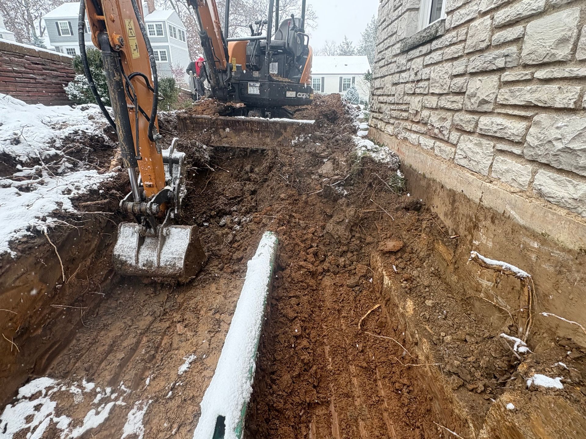 Excavator digging a trench beside a stone house in a snowy yard.